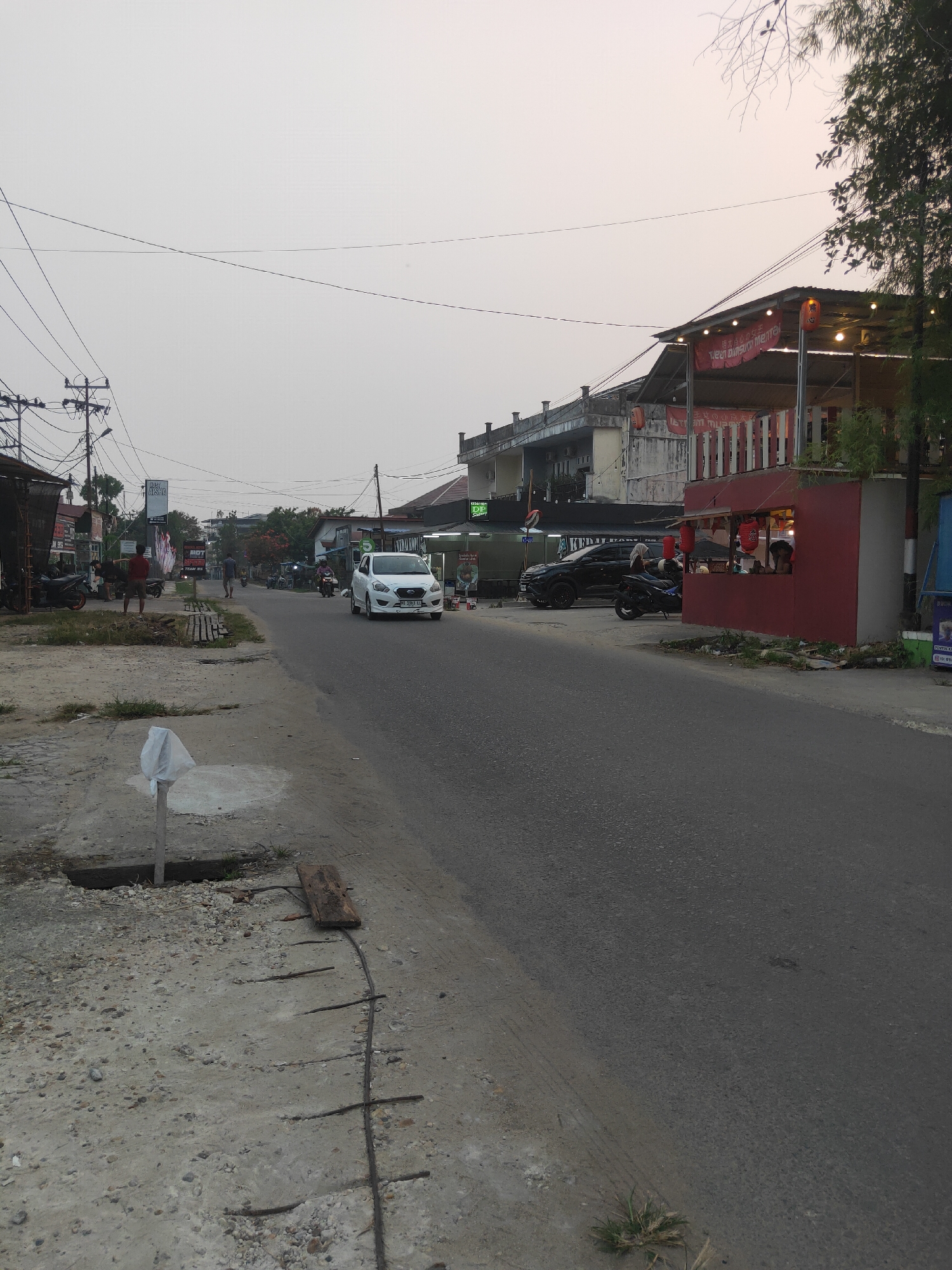 The image shows a street scene in Sintang, Indonesia, at dusk. The street is paved with asphalt and has a concrete shoulder on the left side. Several buildings line the street, including a multi-story building with a red awning and a smaller red food stall. A white car is driving down the street, and several motorcycles are parked on the side. There are power lines crisscrossing the sky, and trees are visible in the background. The sky is a pale orange and gray, indicating the sun is setting. A person is standing on the side of the road, and another person appears to be working at the food stall. The overall impression is one of a typical street scene in a Southeast Asian city.