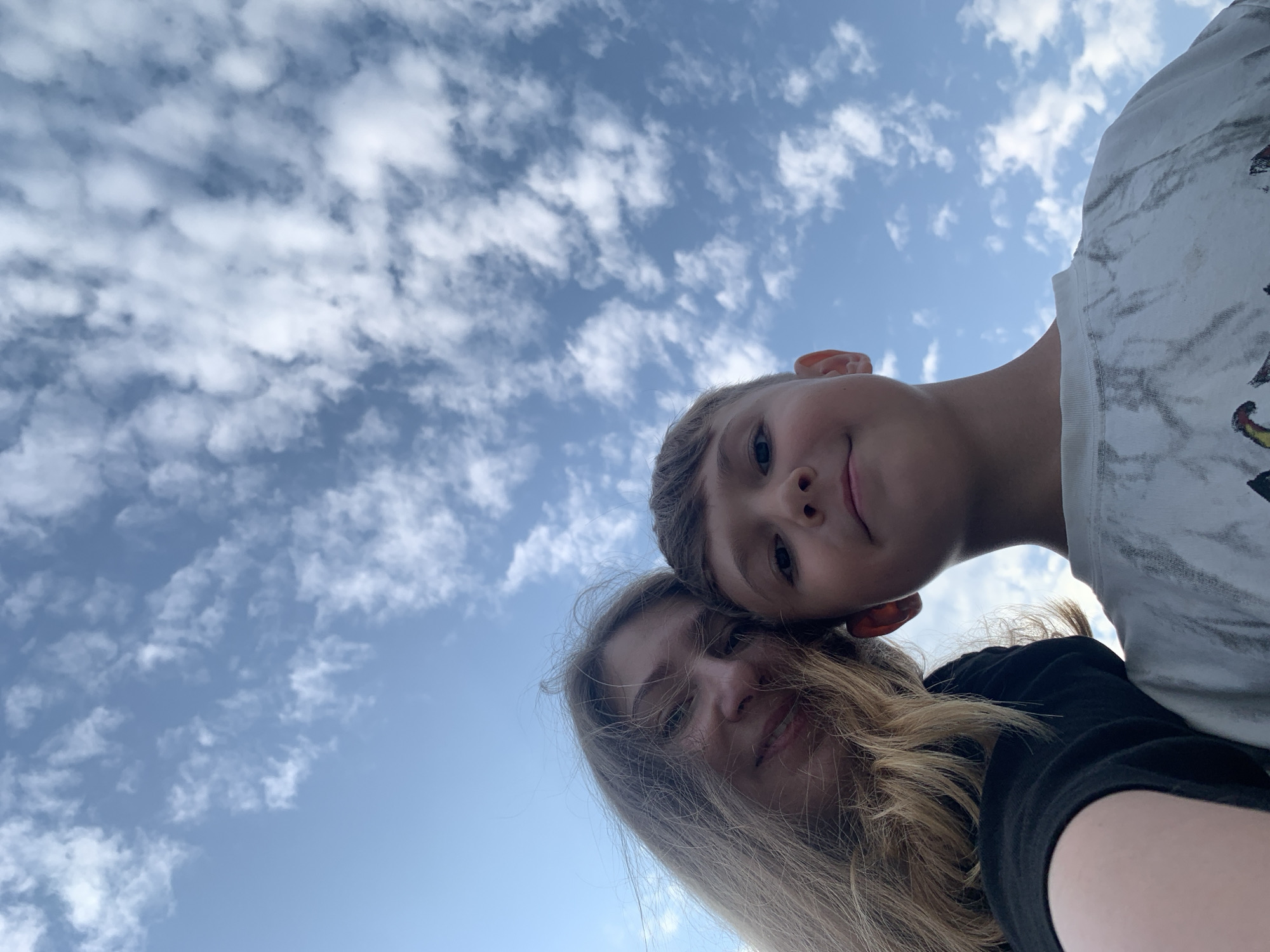 A low-angle, close-up, outdoor selfie shows two individuals, a woman with blonde hair and a young boy, against a backdrop of a blue sky with scattered white clouds. The woman is on the left side of the frame, her face turned slightly towards the camera, with her blonde hair flowing. She is wearing a black top. The boy is positioned to the right of the woman, his face also directed towards the camera. He has short brown hair and is wearing a light-colored t-shirt with a dark, possibly marbled or tie-dye pattern. Both individuals appear to be smiling. The image was taken in Chaikovsky, Russia.