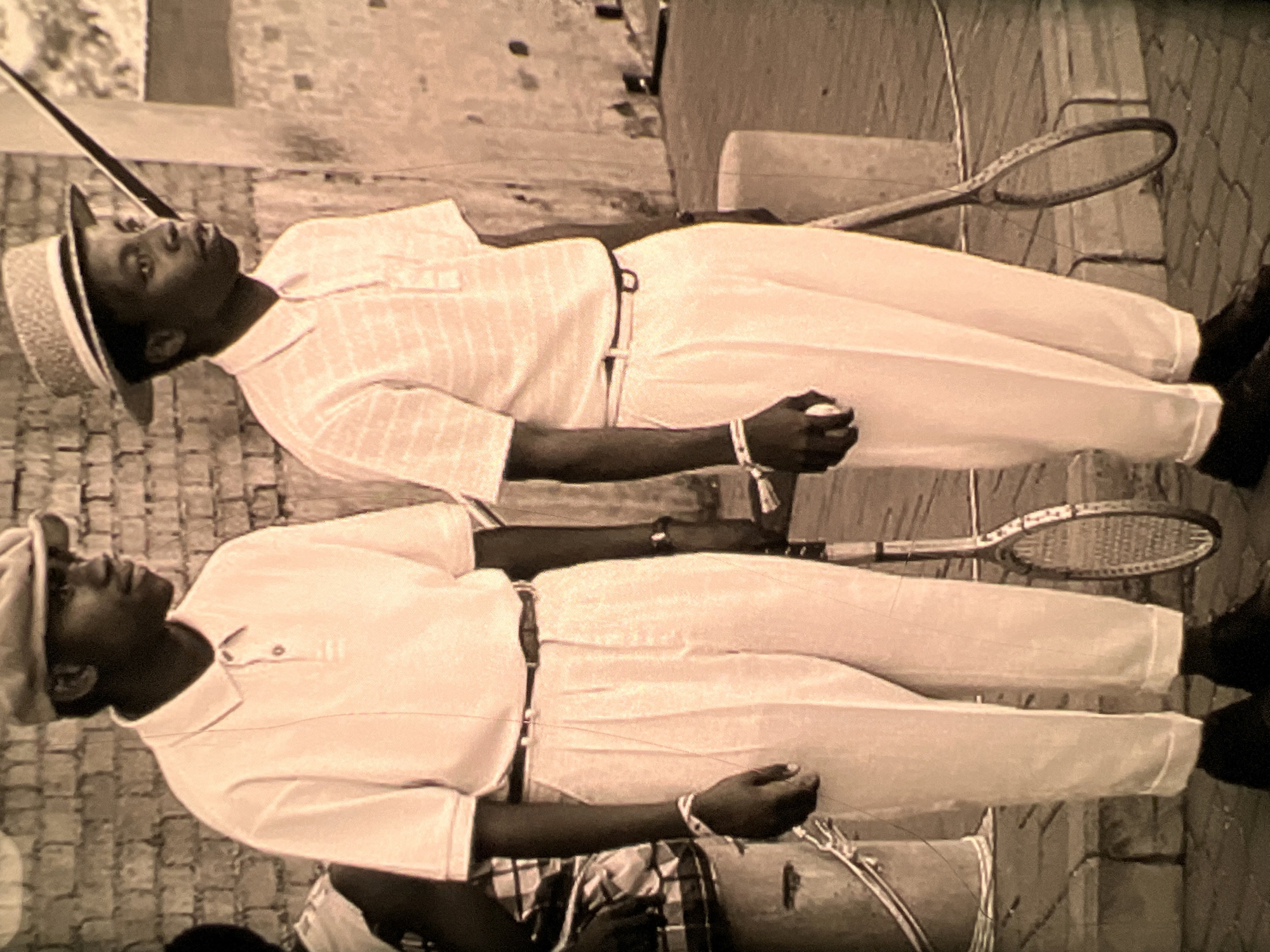 Two young Black men stand outdoors, posing with tennis rackets. The man on the left wears a light-colored polo shirt and trousers, a dark belt, and a flat cap. The man on the right wears a striped polo shirt and trousers, a dark belt, and a straw hat. Both men have beaded bracelets on their wrists. A tennis racket leans against a post behind the man on the right. In the background, a brick wall and what appears to be a bucket are visible. The scene suggests an outdoor setting, possibly a tennis court or recreational area, in Lagos, Nigeria.