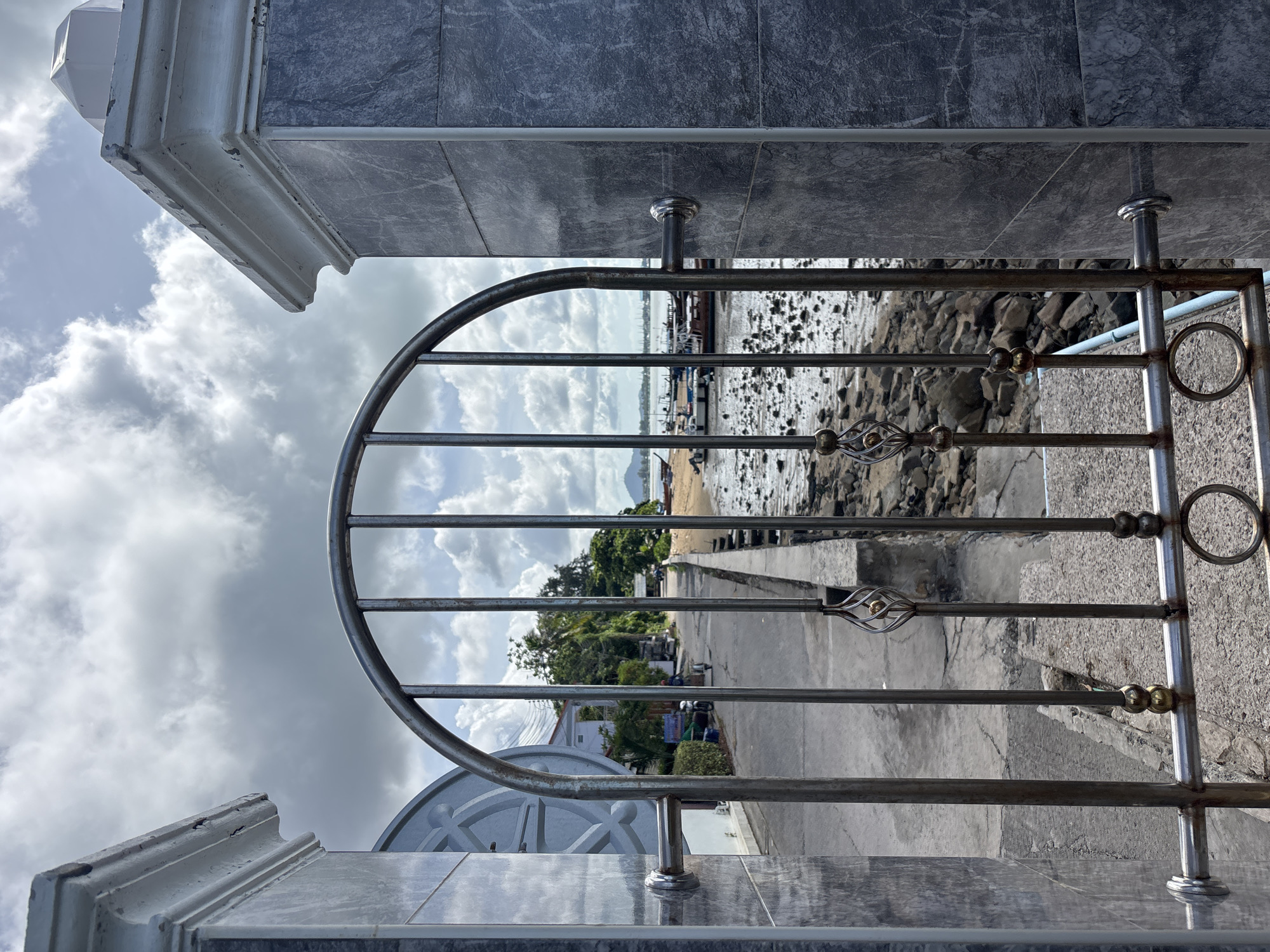 This image, taken in Rawai, Thailand, depicts an open metal gate leading to a street and a beach. The gate is made of vertical metal bars with decorative elements and horizontal bars. Through the gate, a paved street is visible, lined with houses and trees on the left. On the right, a rocky beach is present, with the sea beyond. Several boats are moored in the water. The sky is cloudy, with patches of blue sky visible. The gate appears to be part of a property bordering the street and the beach. The scene suggests a coastal residential area with typical boat activity.