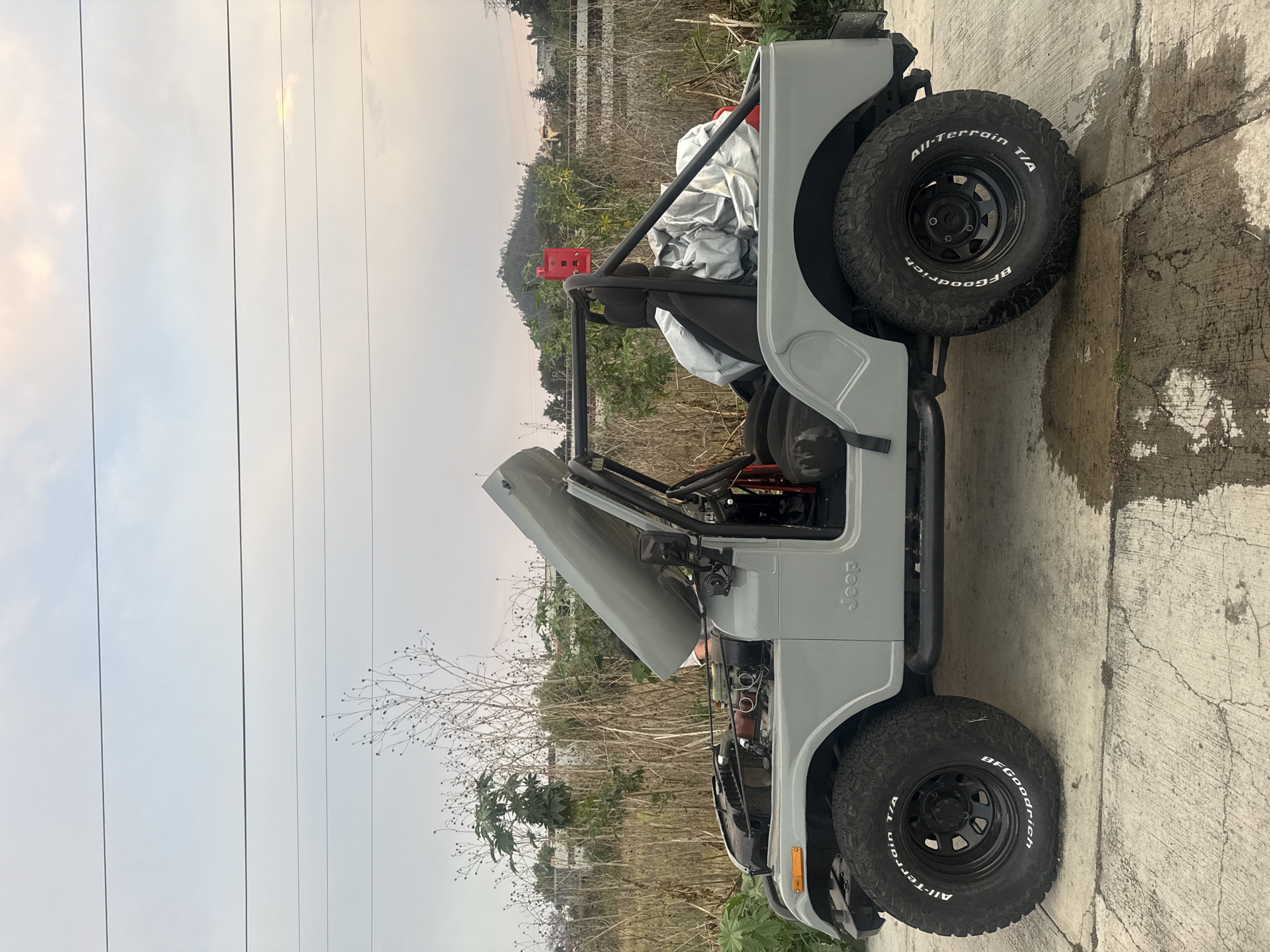 The image shows a grey Jeep parked on a concrete surface, possibly a driveway or road. The Jeep has its hood open, suggesting maintenance or inspection is underway. It's equipped with black wheels and BFGoodrich All-Terrain T/A tires. The background features a mix of vegetation, including trees and shrubs, with an open field or hillside further away. Power lines are visible against the sky. A red object is also visible on the roll cage behind the driver seat. The environment appears to be rural, but it is not possible to confirm the location.