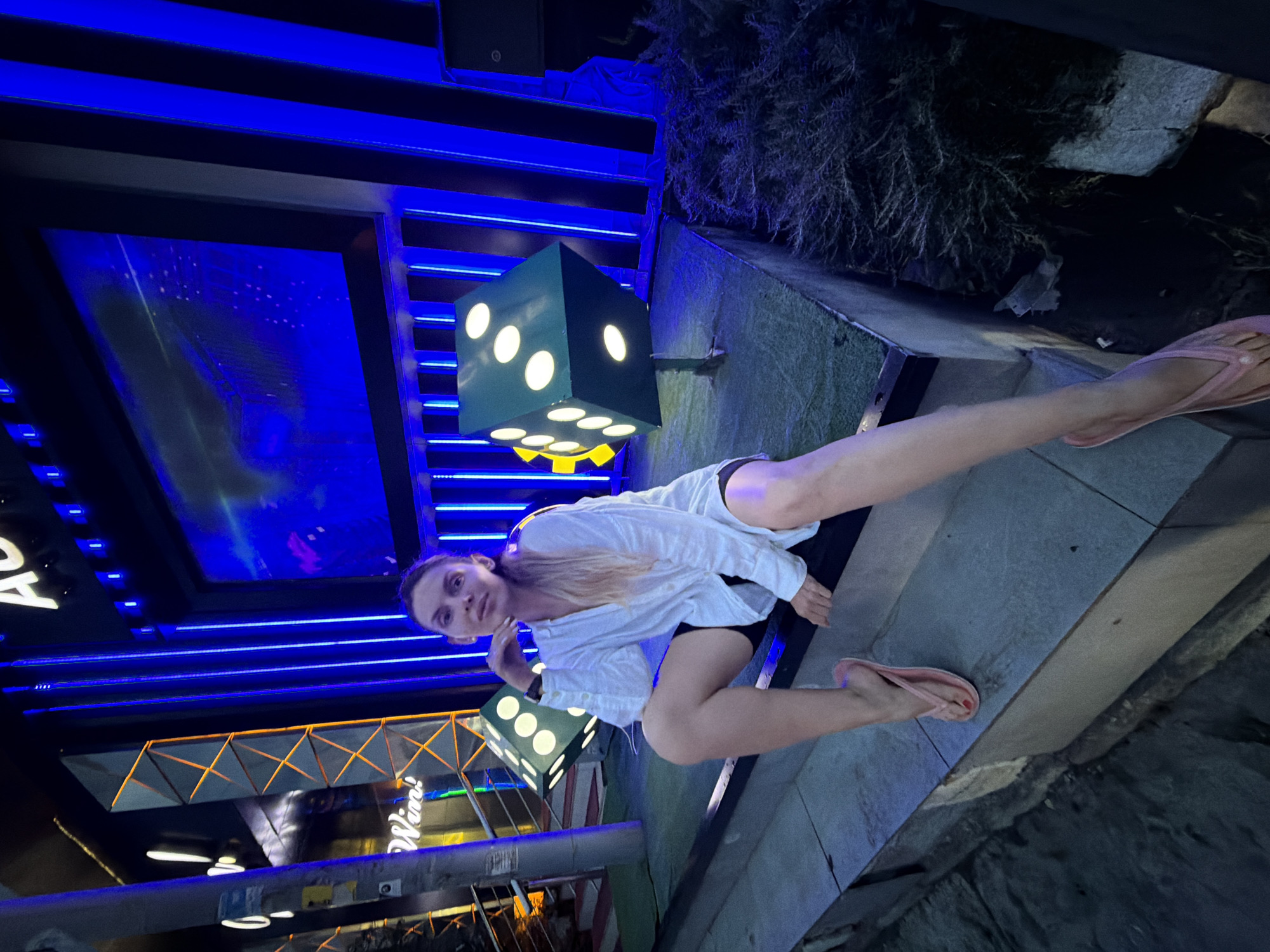 In Budva, Montenegro, a woman with blonde hair pulled back in a ponytail sits on a concrete ledge at night. She is wearing a loose white button-down shirt over black shorts and pink flip-flops. Her right arm rests on her leg, and her left hand is propped under her chin. Behind her, two large, illuminated dice decorations are visible, with the one on the right showing six dots and the one on the left showing five dots. The background features a large screen displaying a blurry image, framed by bright blue vertical lights and some orange geometric designs. The overall scene is set against a dark, urban backdrop, likely a street or entertainment area.