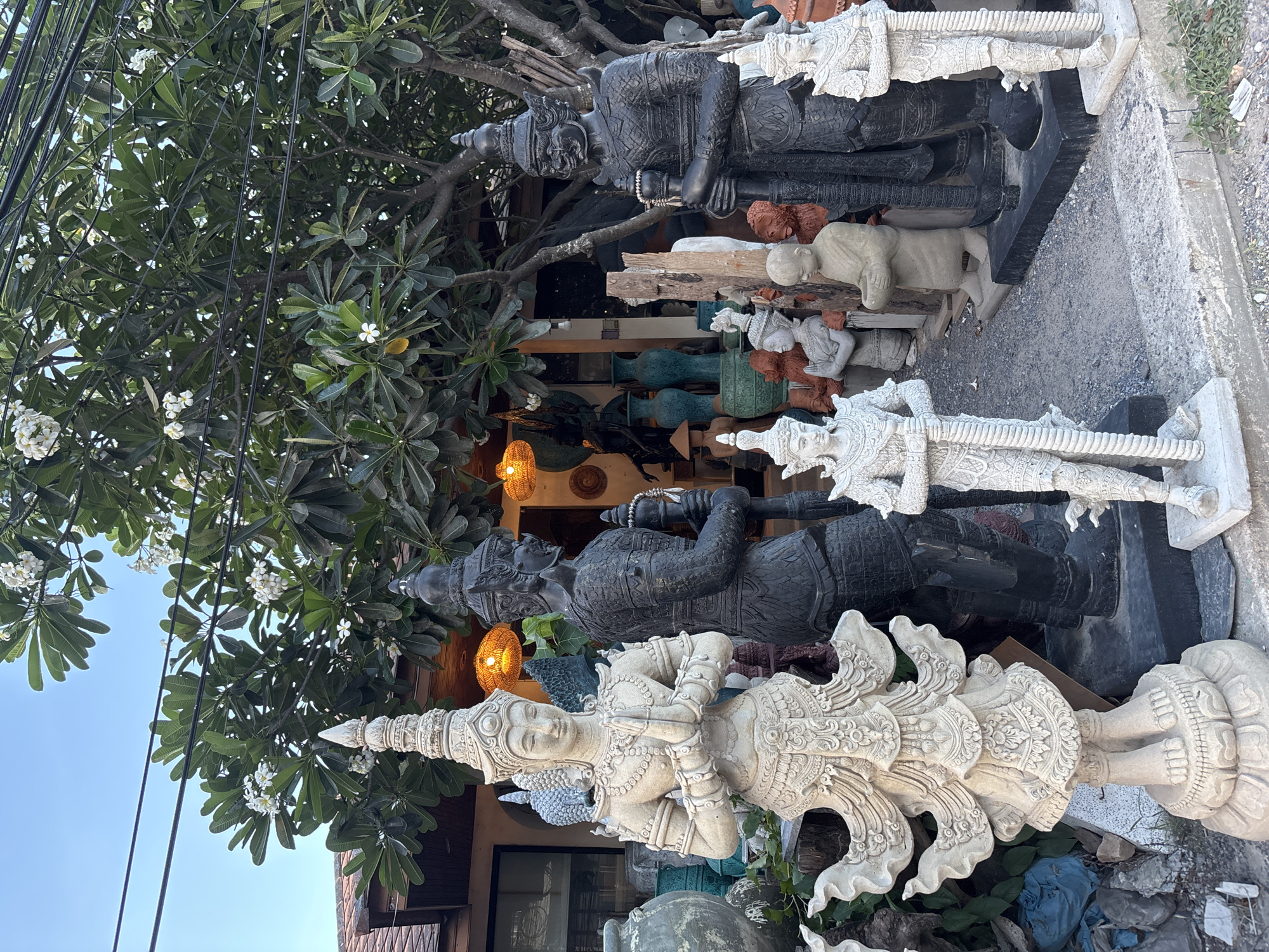 The image shows a selection of stone statues displayed outside a shop in Baan Bo Phut, Thailand. The statues, rendered in white and grey stone, are of various sizes and depict traditional Thai figures. Some are highly ornamented with intricate details. The statues stand on a gravel or paved surface, and a lush green tree with white flowers overhangs the scene. Overhead, there are several visible electrical wires. In the background, part of the shop is visible, with its door and various decorative items like vases and light fixtures.