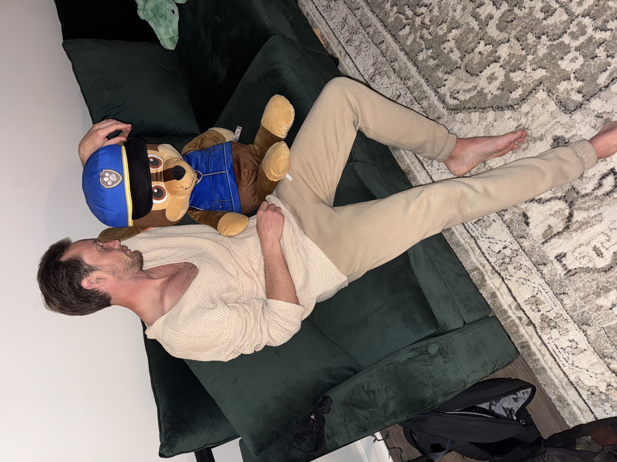 A man is lying on a green velvet sofa with a stuffed animal. The man is wearing a beige sweater and sweatpants. The stuffed animal appears to be a Paw Patrol character named Chase. A patterned rug is on the floor next to the sofa. A black bag is on the floor to the lower right of the sofa. The location appears to be inside a home. The city and state are unknown.