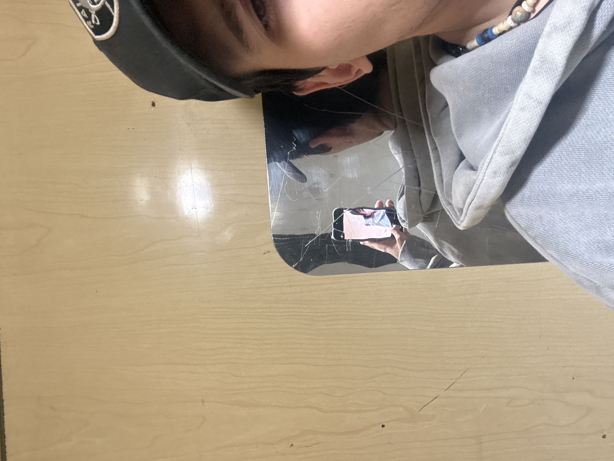 A person wearing a black baseball cap, light blue denim jacket, and beaded necklace is taking a photo in a cracked mirror. The surface beneath the mirror appears to be a light wood. The location is not discernible from the image.
