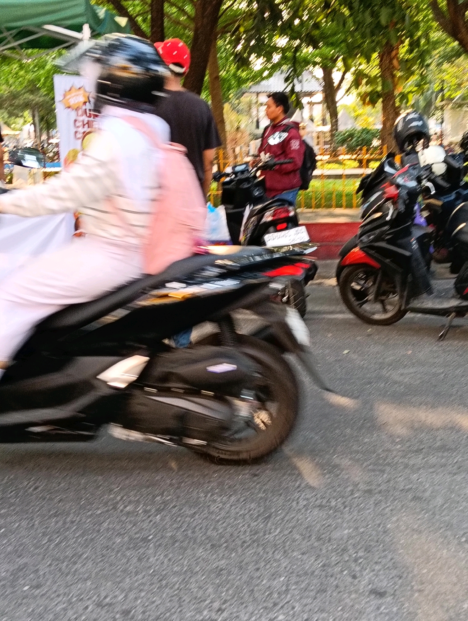 In Karanganyar, Indonesia, this image captures a street scene where several motorcycles are parked or in motion. The primary focus is on a person riding a motorcycle, wearing a black helmet and a light-colored jacket over a pink backpack. To the left, another person wearing a red hat stands near a display booth with a white banner. Further in the background, a person in a maroon jacket is seen on a motorcycle. The environment suggests a public area with trees and a building in the distance. The lighting indicates it is daytime, possibly late afternoon, given the shadows. The image has a motion blur, suggesting movement within the scene.