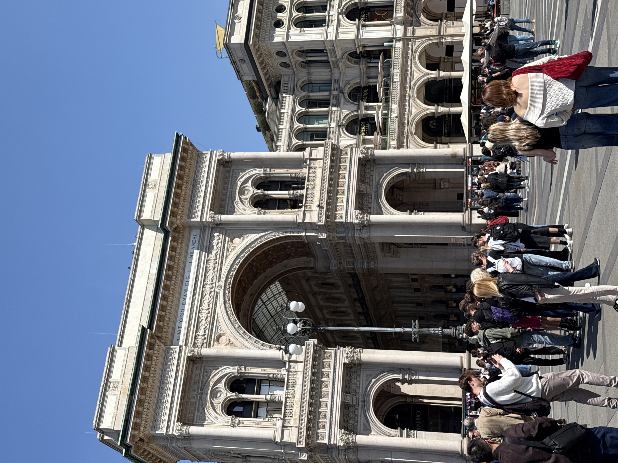 The image features the Galleria Vittorio Emanuele II in Milan, Italy, a renowned shopping arcade. The scene includes the ornate architectural details of the Galleria, with its arches, columns, and decorative carvings. A crowd of people is present, some walking and others observing the surroundings or taking pictures. The image is taken during the daytime under a clear blue sky. The presence of people suggests the location is a popular tourist attraction.
