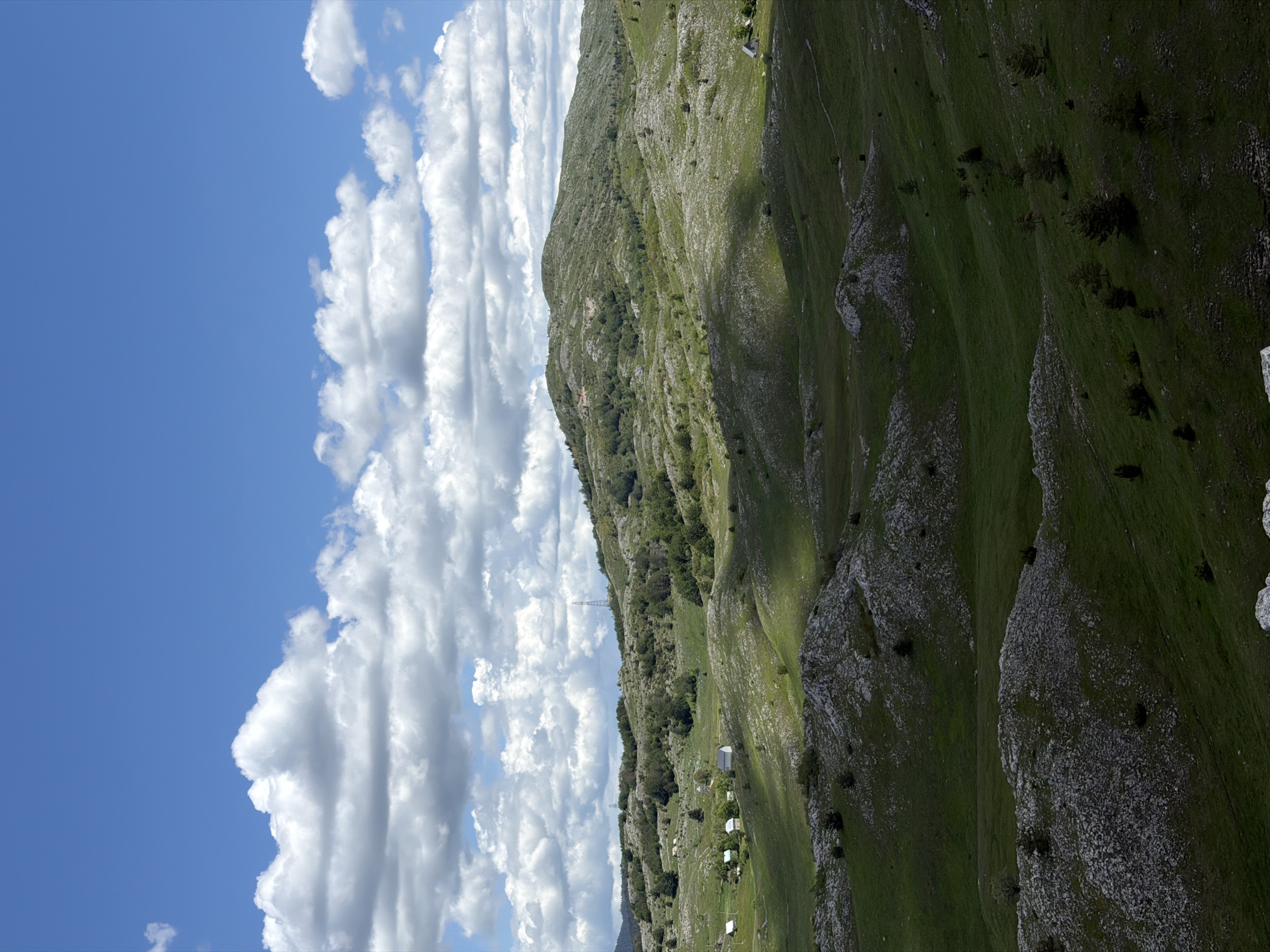 This image captures a wide-angle view of a rural, hilly landscape in Novakovići, Montenegro, under a partly cloudy sky. The upper portion of the frame features a bright blue sky dominated by numerous white and grey cumulus clouds. The lower two-thirds of the image depict an undulating terrain composed of green grassy slopes interspersed with abundant exposed grey and white rock formations, characteristic of a karst landscape. Small, scattered green bushes and sparse trees are visible throughout the hills, with denser tree clusters on some slopes.
In the mid-ground, a small cluster of approximately five to six single-story white-roofed buildings or houses is situated within a shallow valley on the left side. A solitary similar white-roofed building is visible on a distant green slope in the mid-ground on the right. Further into the distance, atop prominent hilltops, at least two tall, lattice-structured metallic towers, resembling communication or power transmission pylons, are present. The scene is illuminated by direct sunlight, creating distinct patterns of light and shadow across the hills, indicating a high sun position. No human figures or discernible animal life are present within the frame.