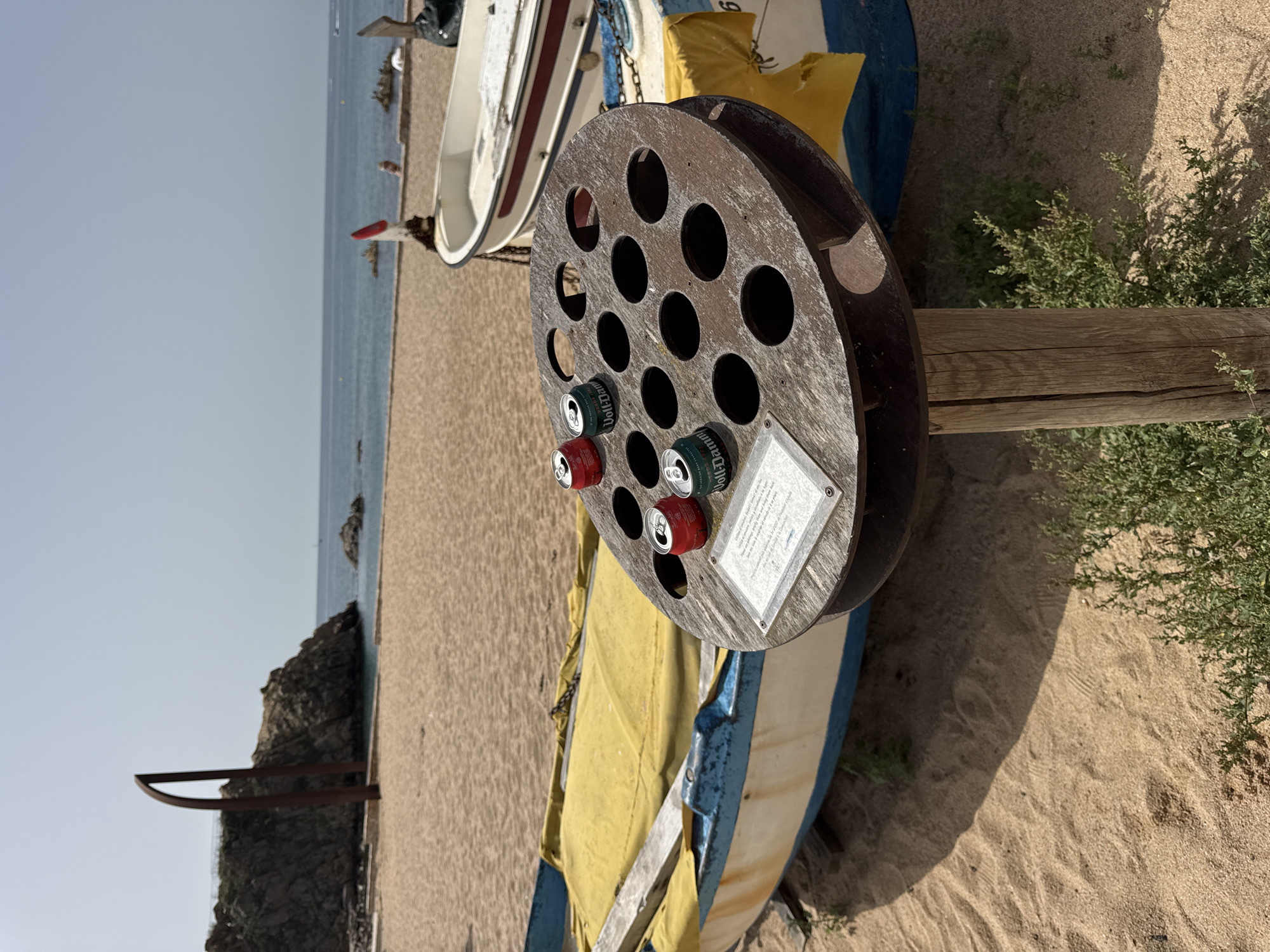 The image depicts a beach scene in Blanes, Spain, with a focus on a circular metal receptacle with numerous holes, possibly a recycled art installation or a unique trash can. Three aluminum cans are placed in some of the holes: two are red and white, and one is green and white. A sign is affixed to the front of the receptacle.
In the foreground, a weathered wooden post supports the receptacle, which is situated on a sandy beach. Two boats are visible behind the receptacle. The boat in the immediate background is partially covered with a yellow tarp and has blue and white hull markings. Another white boat with a red stripe is positioned further back.
The background reveals a calm blue ocean meeting the sandy shore, with a rocky outcrop on the left side of the frame. The sky is clear and bright. The overall atmosphere suggests a sunny day at the coast.