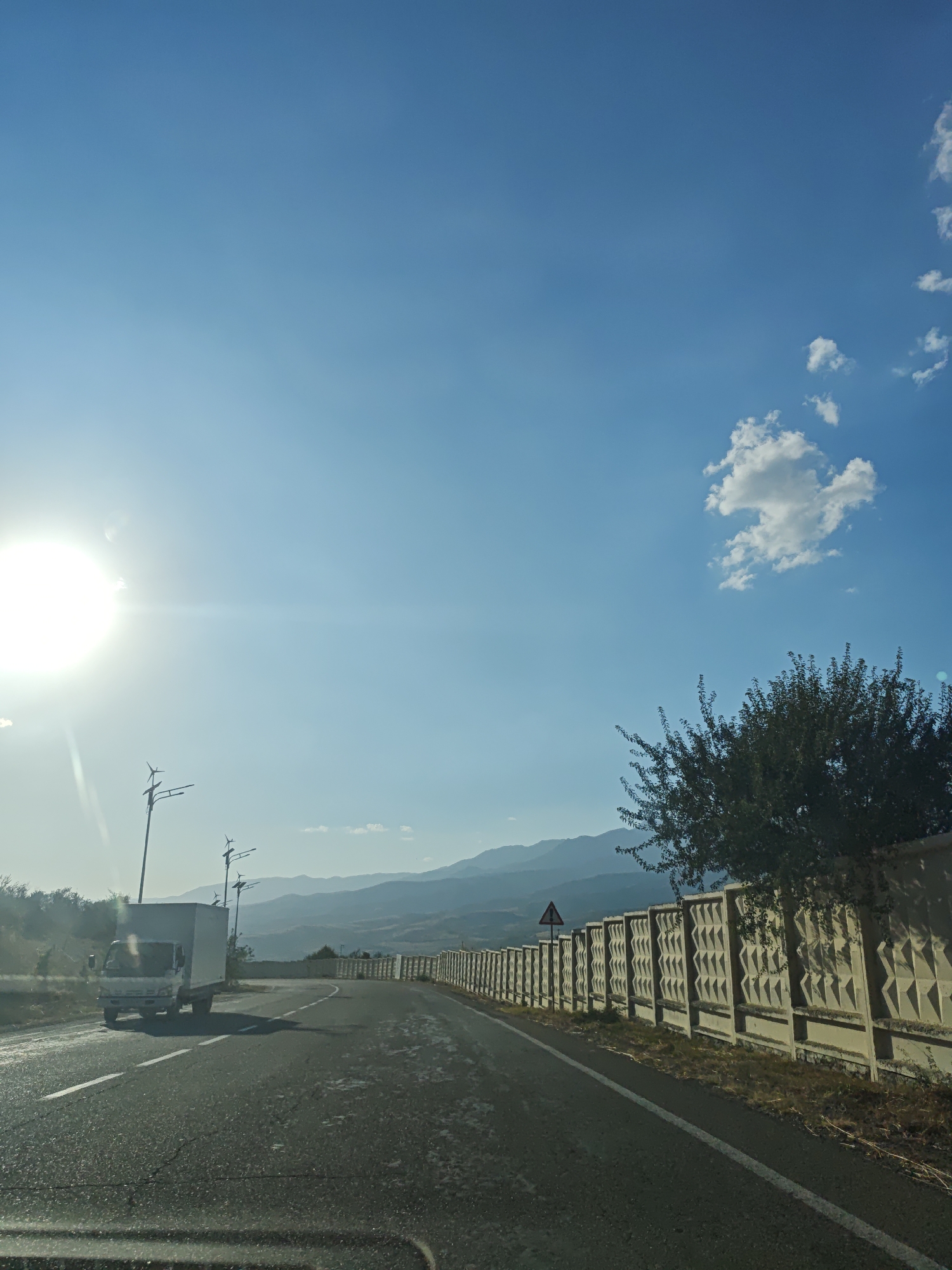 The image captures a view from a vehicle on a paved road, likely in or near Chinarkent, Uzbekistan. The road curves to the right, bordered by a light-colored concrete fence with a diamond pattern. A mature tree with dense foliage overhangs the fence on the right side of the image. In the distance, rolling hills and mountains are visible under a clear blue sky with a few scattered white clouds.
To the left of the road, a white box truck is traveling in the opposite direction. Beyond the truck, several lampposts with what appear to be small wind turbines at their apex are visible. The sun is a bright, overpowering presence in the upper left quadrant of the sky, creating lens flare and washing out some of the details in that area. The overall environment appears dry and somewhat arid, consistent with the geography of Uzbekistan. There are no visible people in the image. The scene is captured during daylight hours.