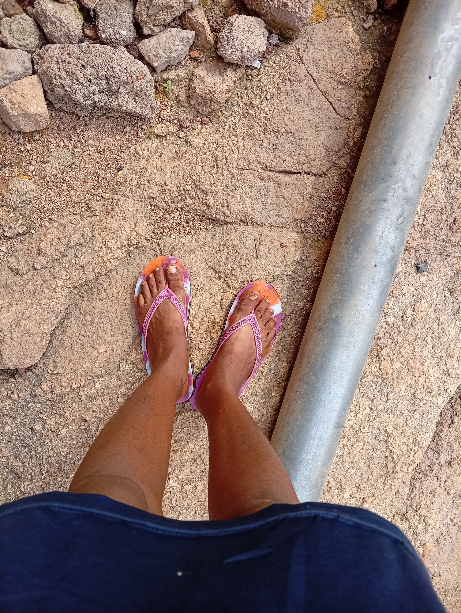 The image shows a person's legs and feet wearing flip-flops, standing on a rocky, earthy surface. The flip-flops are pink with an orange and white tie-dye pattern. The person is wearing dark blue shorts or a skirt. To the right of the legs is a large, weathered metal pipe running diagonally across the frame. The ground is composed of large, rough stones and smaller pebbles. In the upper left of the image, more rocks and dirt are visible, suggesting an outdoor, unpaved setting. The lighting suggests it is daytime. The scene is likely outdoors in Jos, Nigeria, given the rocky terrain and general environment. There is no discernible text in the image.