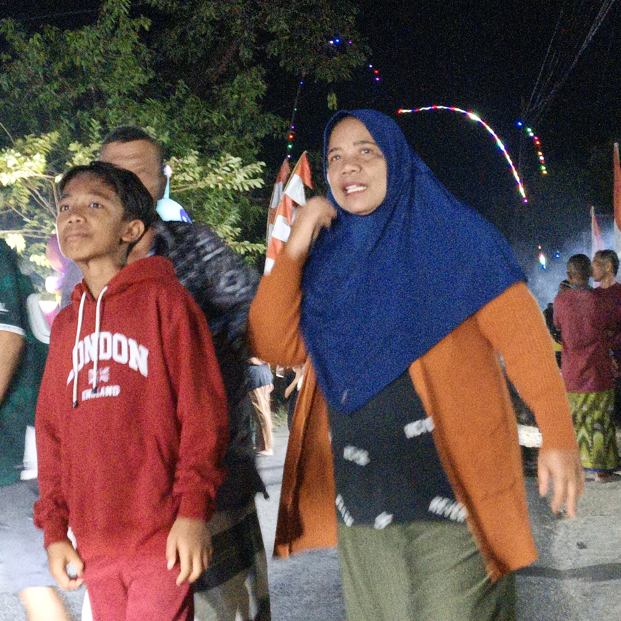 In the city of Kalitorong, Indonesia, at night, a group of people are gathered outdoors. The foreground features a young boy wearing a red hooded sweatshirt with "LONDON ENGLAND" printed on it, and he is looking to his left. To his right, a woman is wearing a blue hijab and an orange sweater. Behind them, other individuals are visible, and in the background, decorative lights are strung across the scene, casting a festive glow. The ground appears to be a paved surface, likely a street or pathway, and trees with dense foliage are present. The overall atmosphere suggests a community event or celebration.