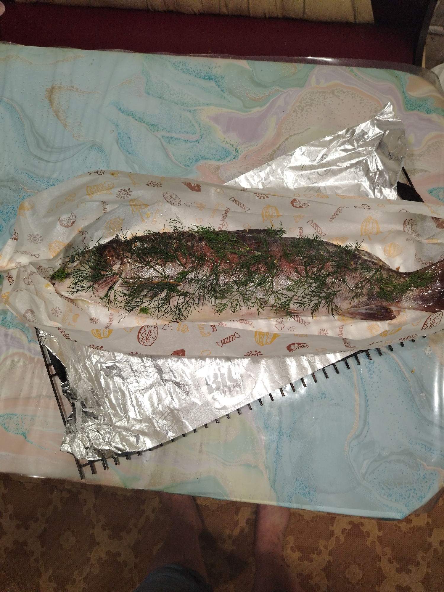 A whole, raw fish is laid out on a patterned paper liner, which is then placed on a shiny, crinkled aluminum foil. The fish is covered in fresh dill, with its scales and fins visible. The paper liner has an assortment of printed images, including what appear to be baked goods like donuts and croissants, and possibly some text in Russian.
The scene is set on a table covered with a blue and white swirled, marbled pattern tablecloth. In the background, a dark red cushioned surface is visible. To the left of the fish, the edge of a metal rack with small, evenly spaced teeth is visible, partially covered by the aluminum foil. A small portion of a person's bare feet is visible at the bottom of the frame, standing on a brown patterned carpet. The lighting suggests an indoor setting, possibly during the day, with no clear indication of the time of day or weather. The overall impression is of food preparation, with the fish being prepared for cooking. The visible text on the paper liner is not clear enough to be fully deciphered.
The location context of Святогорье, Russia, implies a region known for its natural beauty, though this is not directly evident from the image. The image focuses on the prepared ingredients and the immediate surroundings.