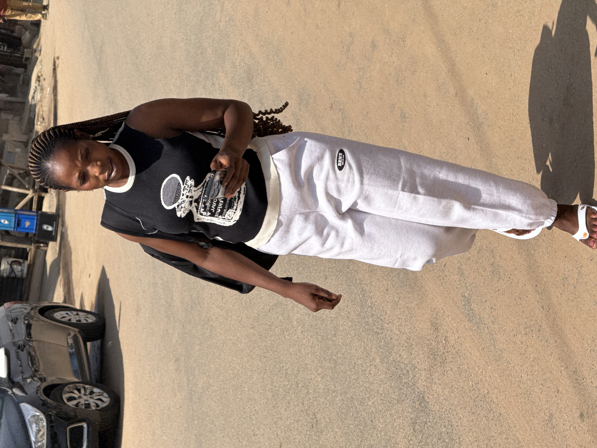 A young woman with braided hair is walking on a dirt road in Abuja, Nigeria. She is wearing a black sleeveless shirt with a white trim and a graphic design on the front, and light grey sweatpants. She is holding a smartphone in her right hand and has a black backpack slung over her left shoulder. The road is paved with a light brown, sandy material, and there are shadows cast on the ground, indicating it is daytime. In the background, a black car is parked on the left, and some blue crates and wooden structures are visible further down the road, suggesting a market or commercial area. A person in a brown shirt and tan pants is visible in the far background on the right.