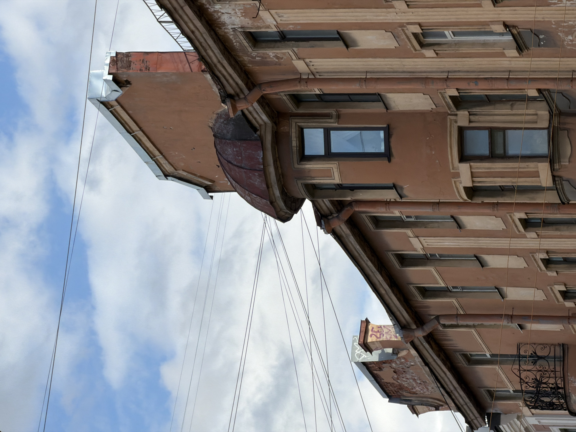 The image shows a section of a building facade in Saint Petersburg, Russia, viewed from a low angle looking up. The building is brown with architectural details around the windows and along the roofline. There are several windows visible, some with black frames and glass reflecting the sky. Gutters and drainpipes run along the side of the building. The sky is visible in the background, with white and gray clouds. Power lines or communication cables are strung across the sky in the foreground.