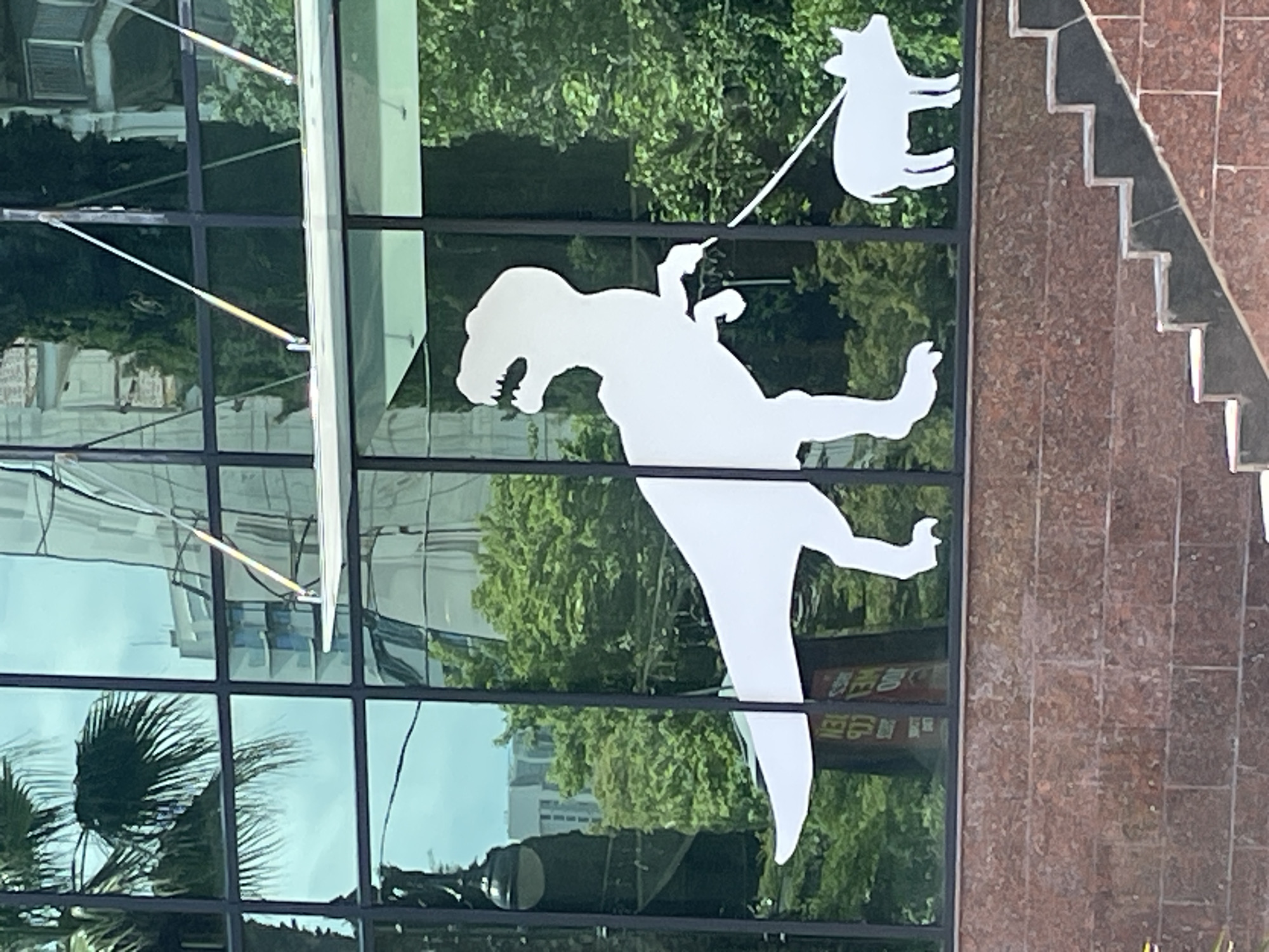 This is a photograph taken on a cloudy day of a building's reflective glass facade with decals of a Tyrannosaurus Rex and a pig. The T-Rex decal is large and white, walking to the left while holding a leash with its right hand. The pig decal is smaller and white, walking to the right on the leash. The decals are placed on the glass in a way that they appear to be walking in front of reflections of trees, buildings, and a palm tree. Below the glass facade is a red brick wall. To the right of the wall, a set of stairs with dark treads and red brick risers is visible. The building appears to be located in Sochi, Russia, as suggested by the distinct red brick building material and the architectural style often found in the city.