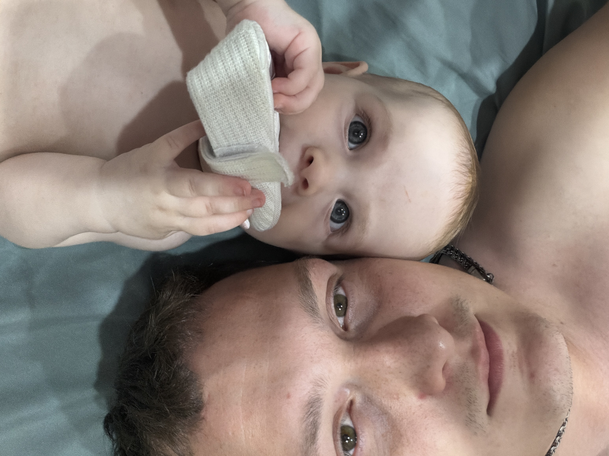 The image shows a close-up, high-angle selfie of a man and a baby lying on a teal-colored surface. The man is on the left, his face filling the lower left quadrant of the frame. He has short, dark brown hair, brown eyes, and light skin with visible pores and some freckles. He is wearing a necklace with a silver chain. The baby is on the right, oriented upside down relative to the man. The baby has fair skin and light blue eyes, and is holding a white, textured object, possibly a sock or a piece of fabric, up to its face. The teal surface appears to be a bed or a mat. The lighting creates soft shadows on the surface. There is no indication of the location in Noviy, Russia.