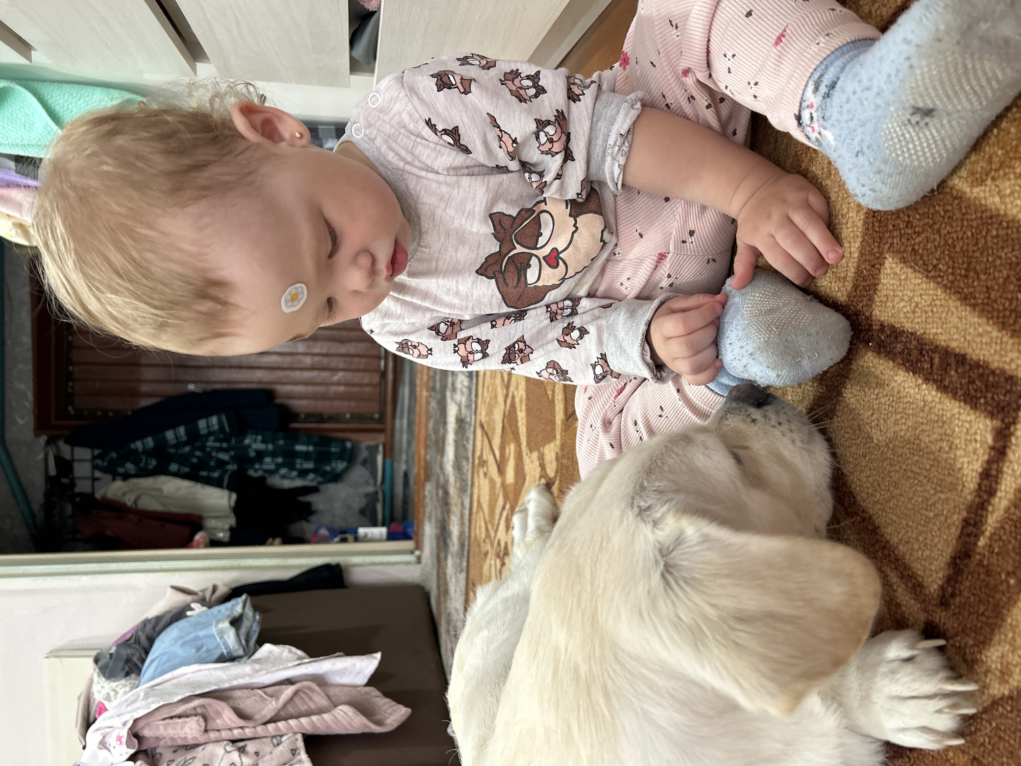 This media file captures an intimate indoor scene in Азинский, Russia, featuring a young child and a light-furred dog. The child, with blonde hair and a small white flower sticker on their forehead, is dressed in light-colored pajamas featuring an owl pattern on the top and pink-striped pants. They wear light blue socks. The child is sitting or lying on a brown patterned rug, looking down at the dog. The dog, which appears to be a puppy or young dog with light fur, is lying close to the child, its head resting near the child's foot. The child's hands are near the dog's paw, suggesting a gentle interaction or observation. In the background, a light-colored dresser or cabinet is visible, along with piles of clothes, indicating a domestic setting. The scene evokes a sense of calm and companionship between the child and the pet. The overall lighting suggests it is daytime indoors. There is no visible text.