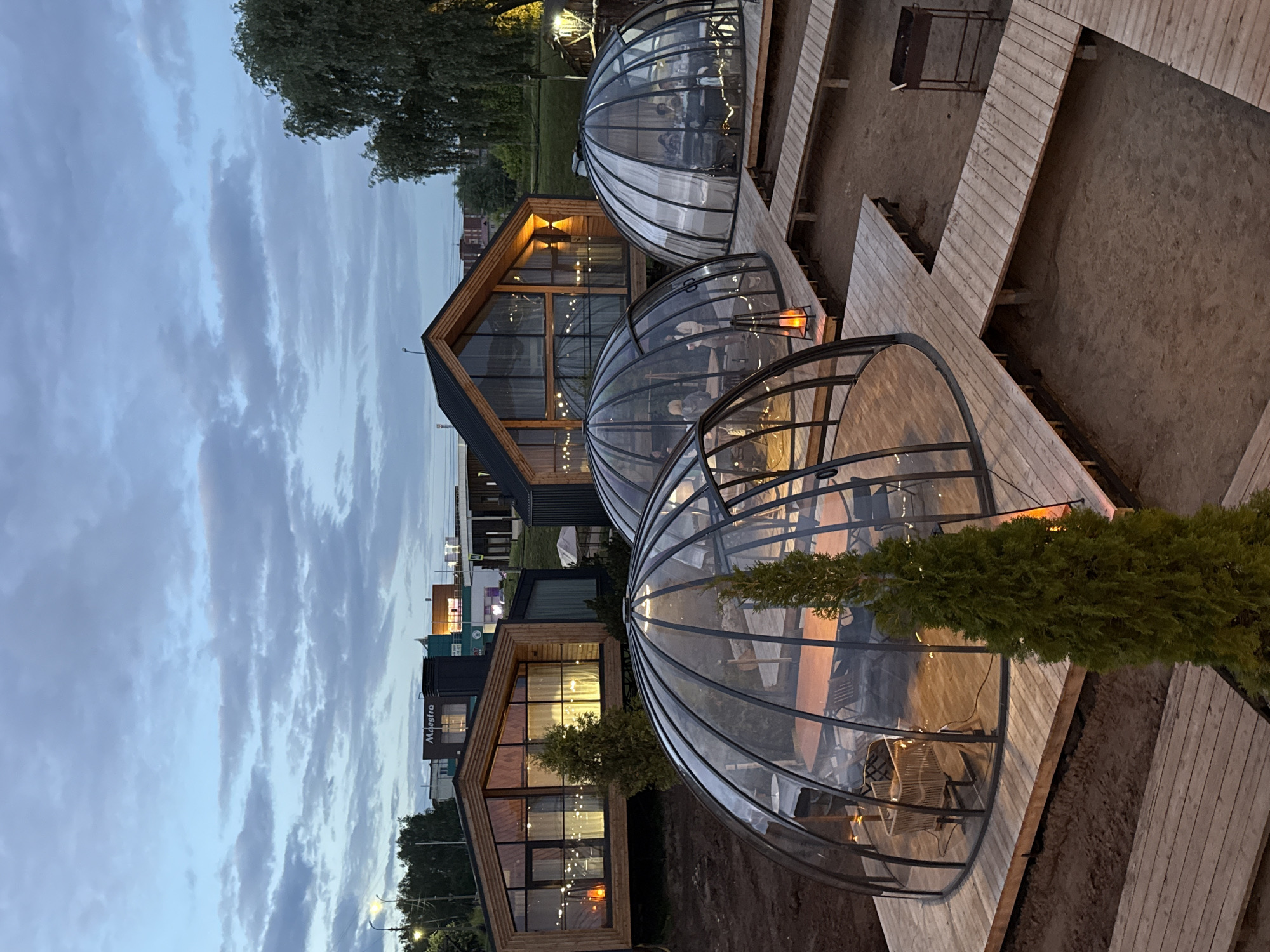 The image depicts an outdoor dining area at dusk in Votkinsk, Russia. The scene features several transparent, geodesic-shaped dining pods arranged on wooden platforms. Inside the pods, patrons are visible, suggesting that dining is taking place. Warm light emanates from within the pods and from string lights strung across the wooden structures, creating a cozy and inviting atmosphere.
Behind the dining pods, a modern building with large windows and a triangular roof is illuminated from within. The building's architecture appears contemporary, with wood and dark metal accents. Signs on the building are partially visible, with one displaying the text "Maestro".
The sky is cloudy, indicating evening or a period of overcast weather. The ground surrounding the wooden platforms is sandy or earthy. The overall impression is one of a relaxed evening gathering in a unique outdoor setting. The lighting suggests that it is either early evening or late twilight.