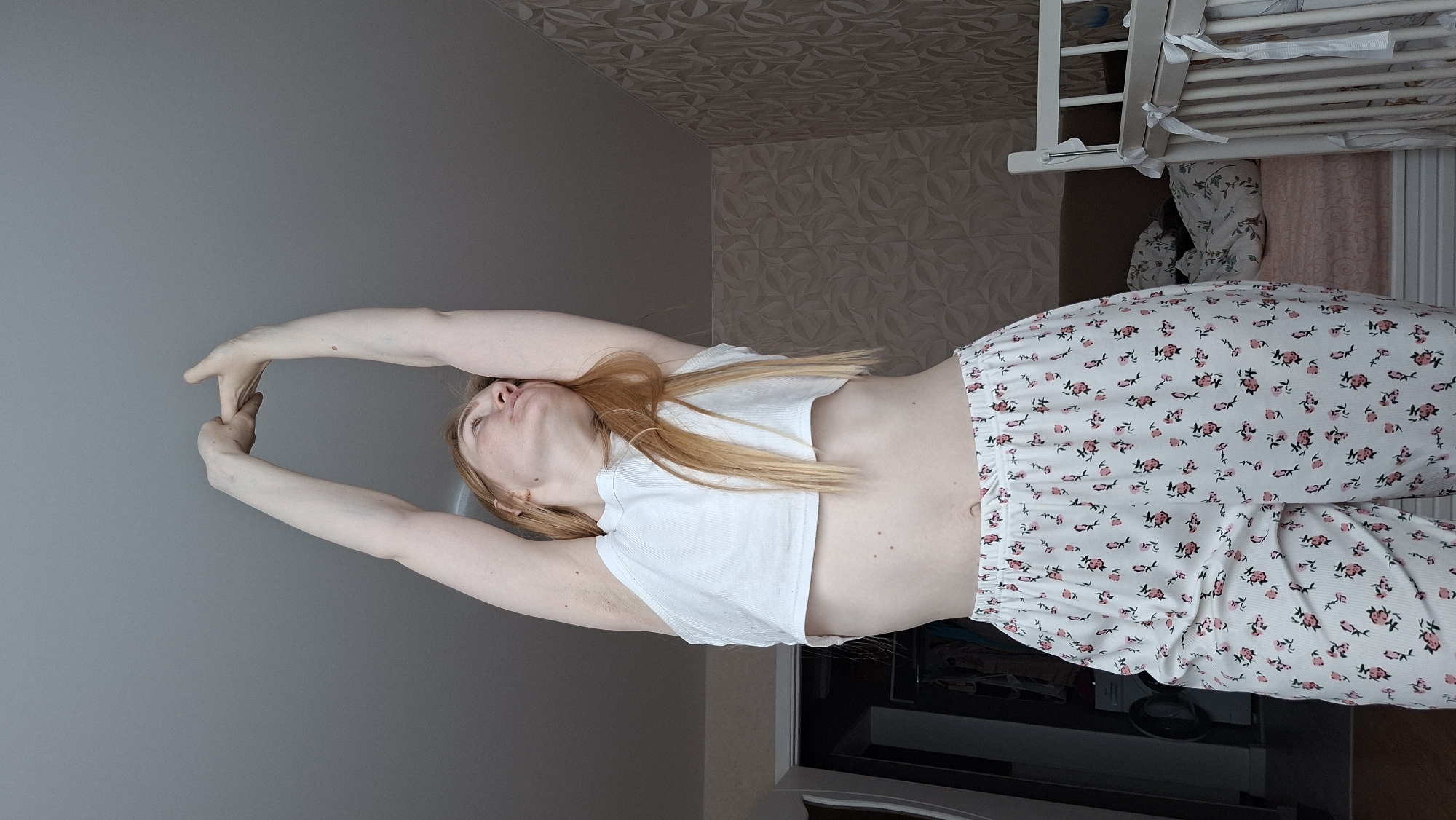 A young, fair-skinned woman with long blonde hair is stretching in a room with patterned wallpaper. She is wearing a white cropped t-shirt and white floral patterned pajama pants. Her arms are raised above her head, and her hands are clasped together. The room includes a grey wall, a partially visible crib, and a dark-colored cabinet. Based on the available information, it is not possible to determine the exact location within Chaikovsky, Russia.
