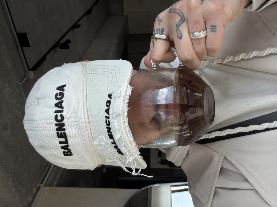A close-up, chest-up shot captures an individual indoors, holding a clear glass mug containing a dark brown liquid, positioned directly in front of their face. The person wears a distressed white baseball cap featuring the black embroidered text "BALENCIAGA" prominently on the front, with a smaller, partially obscured "BALENCIAGA" below. Beneath the cap, a light-colored hoodie with a white and black striped drawstring is visible, layered under a light-colored trench coat. The person's right hand, adorned with a silver-colored ring on the pinky finger, a textured silver-colored ring on the ring finger, and a partially visible silver-colored ring on the middle finger, grips the mug's handle. Tattoos are visible on the hand, including an outlined snake on the back, a small red mark near the thumb, and three vertical lines on the side of the index finger. The background reveals a modern interior with a concrete ceiling featuring black track lighting fixtures. A faint reflection in the mug's liquid shows a sky with clouds, suggesting a window or outdoor view nearby. The image provides no discernible visual information to definitively identify the location as Yerevan, Armenia.