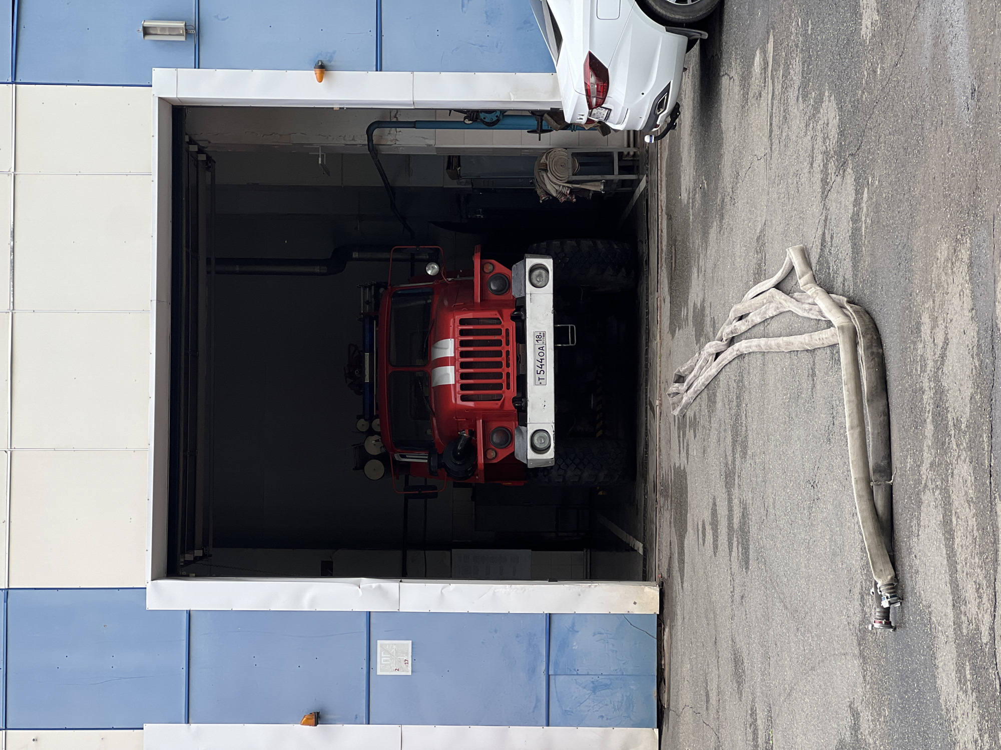 This image captures a fire truck parked inside a bay of a building, likely a fire station, in Izhevsk, Russia. The fire truck is predominantly red with white accents on its grille and sides. Its large tires are visible, suggesting it's an all-terrain vehicle. Above the truck, a white car is partially visible, parked in the same bay. Outside the bay, a coil of thick, grey hose lies on the asphalt ground. The building's exterior is a mix of light-colored panels and blue panels, with a white frame around the bay opening. An orange warning light is visible on the exterior of the building. The ground is asphalt and appears wet in some areas, possibly from recent rain or cleaning. The lighting suggests it is daytime, and the weather appears overcast. A license plate on the fire truck is partially visible with text, including Cyrillic letters and numbers. The overall scene conveys a sense of readiness and preparedness.