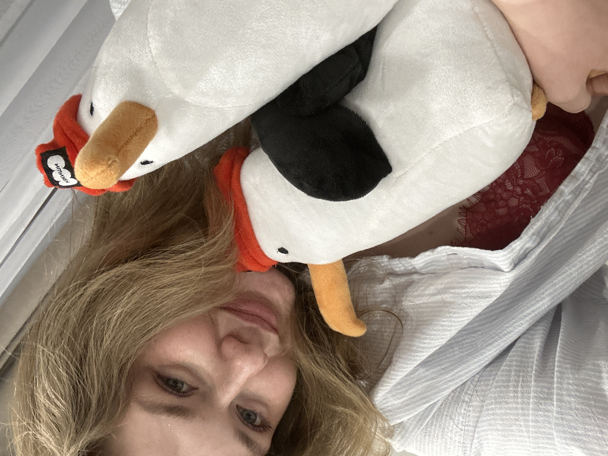 A woman is lying on a bed or surface covered with a white and light blue striped fabric, possibly a shirt or bedding. Two stuffed animals, appearing to be white geese with orange beaks and red necks, are positioned above her head. One of the geese has a black patch on its head. The woman has light blonde hair and blue eyes. A piece of red lace material is visible in the upper right area of the image, slightly obscured by the geese. There is no information in the image that identifies the location as Kazan, Russia.