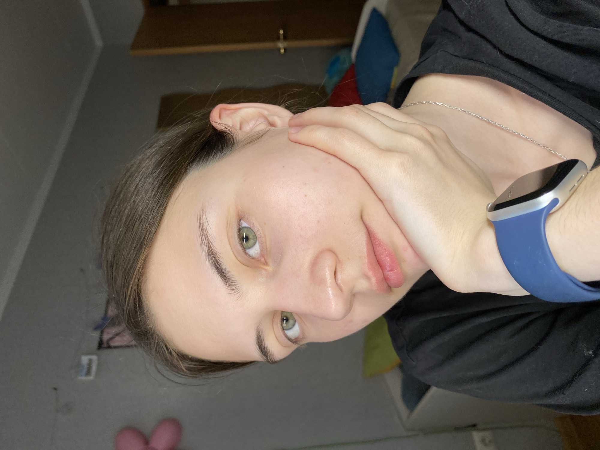 A young woman with fair skin, light brown hair, and green eyes is shown from a close, low-angle perspective. She is wearing a black shirt and a silver necklace. She has a blue Apple Watch on her left wrist. Her left hand is gently touching her face. The background shows a room with a shelf, some colorful items, and a gray wall. There is a pink object on the floor, and some other items are visible against the wall. The location in Votkinsk, Russia cannot be determined from this image.