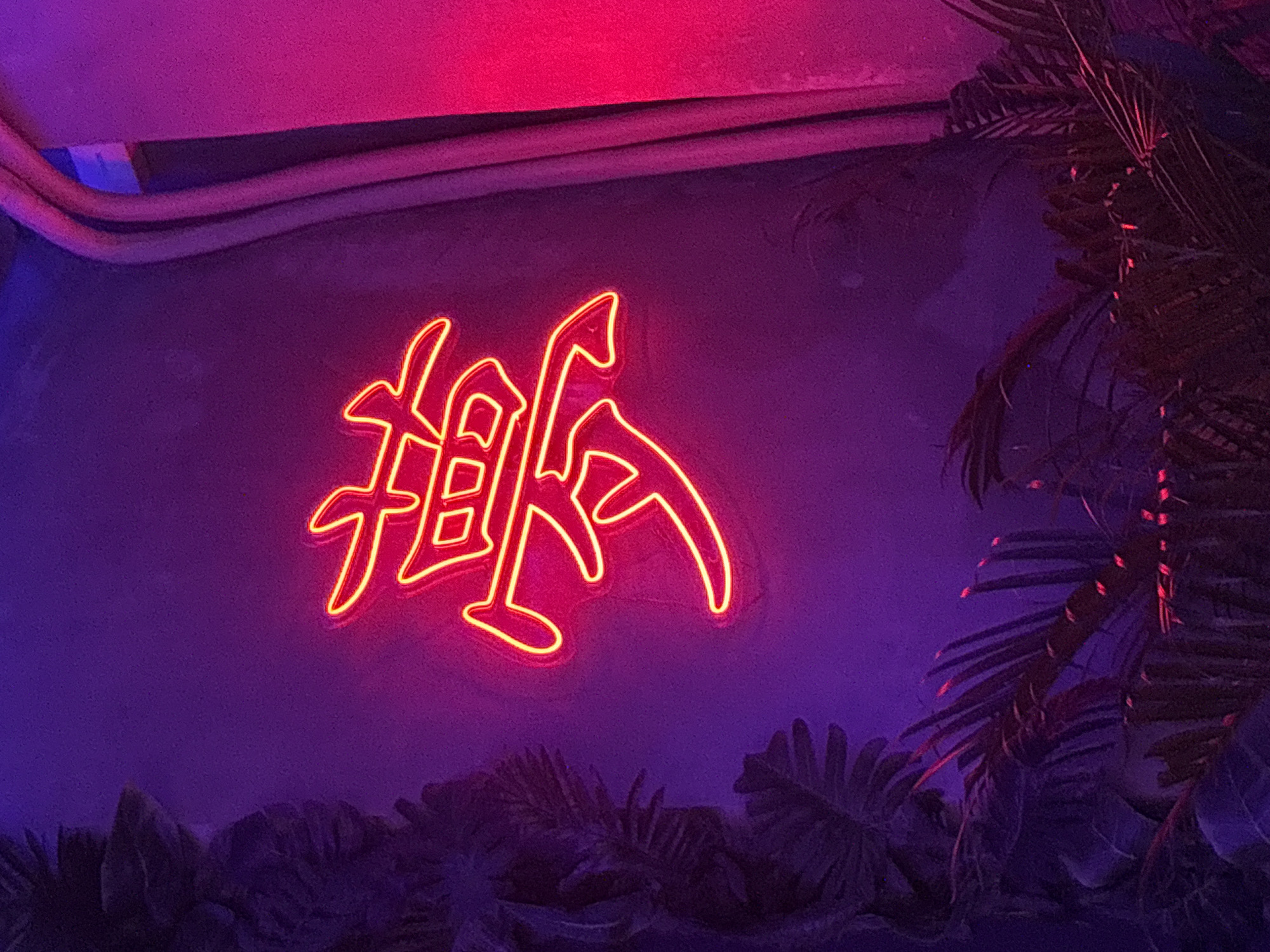 A neon sign depicting a calligraphic-style word in red/orange is affixed to a wall, likely indoors. The wall is a textured, matte purple color. The sign is positioned to the left of a palm tree, the leaves of which frame the right side of the image. Below the sign and palm tree is a dark-colored arrangement of what appear to be other tropical plants. The lighting suggests the scene is in a dim environment with a purple or reddish light cast. Above the sign are parallel pipes or conduits attached to the wall. The image appears to be taken in Kuta Selatan, Indonesia, but no specific details confirm this location from this photograph.