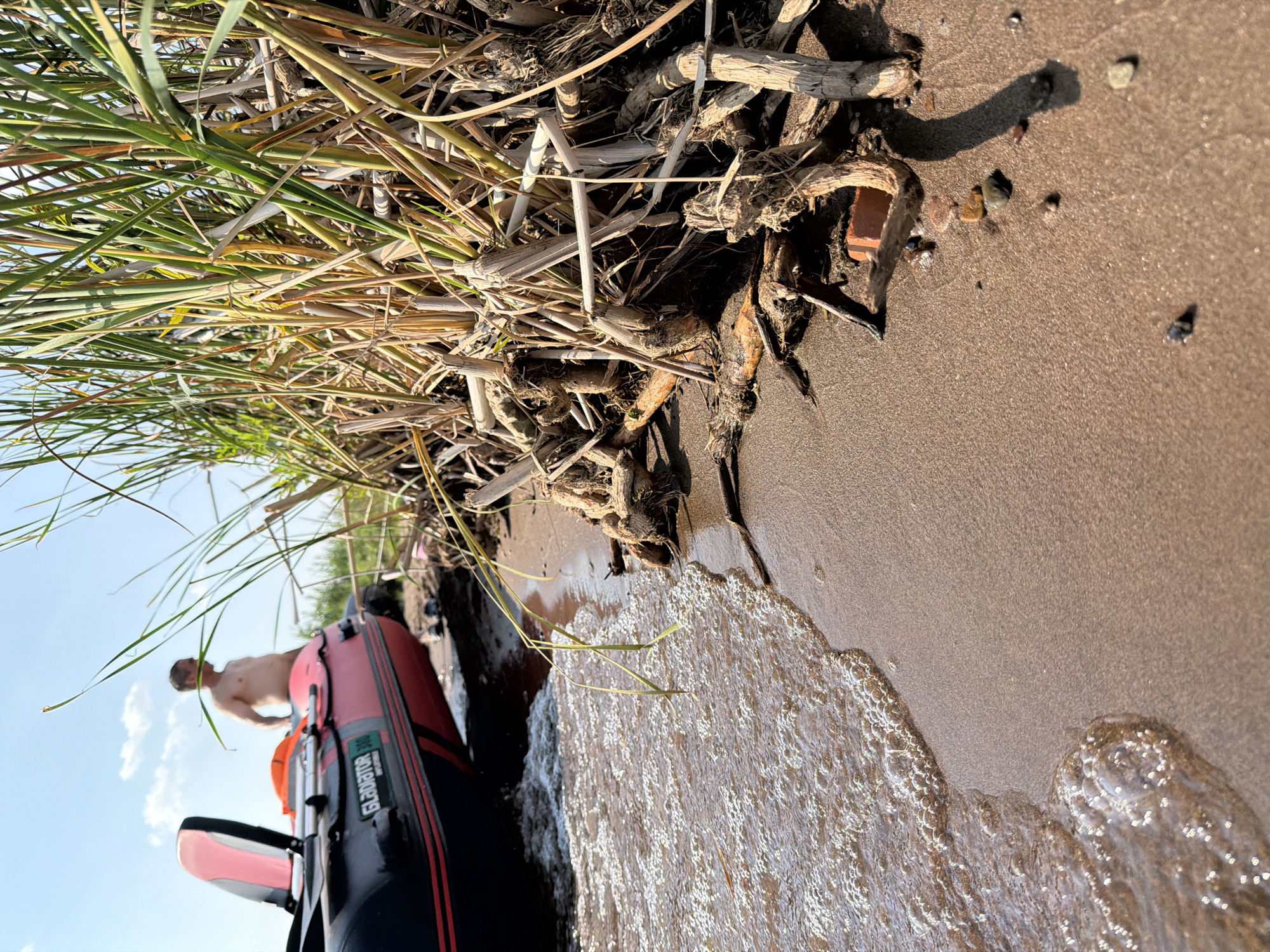 An unclothed man stands near an inflatable boat on a sandy shore. The boat is red and black with the brand name "Gladiator" visible. Behind the boat, dense, dry reeds and driftwood are piled on the sand. The water's edge shows gentle waves lapping onto the shore. The sky is blue with scattered clouds. The scene appears to be a natural outdoor setting, possibly a riverbank or lake shore. The location is identified as Krasnyi Bor, Russia.
