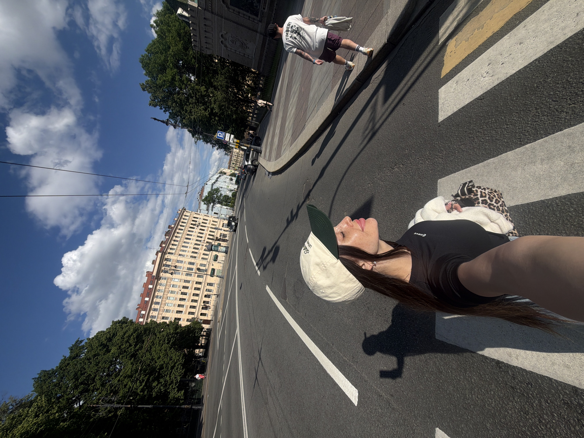 The image captures a street scene in Saint Petersburg, Russia, likely Nevsky Prospekt, given the architecture and layout. In the foreground, a woman is taking a selfie on a pedestrian crossing. She is wearing a black t-shirt, a beige baseball cap with a green brim, and appears to have a leopard-print bag draped over her shoulder. Her face is turned to the side, with her eyes closed, enjoying the sunlight. Her shadow is cast on the asphalt behind her.
In the background, a wide street with multiple lanes of traffic is visible. Buildings with classical architecture line the street, featuring multiple stories and rows of windows. Mature trees with lush green foliage flank the street, casting long shadows. Several cars are on the road, and a few pedestrians are walking on the sidewalk. One man in a white t-shirt and shorts is walking away from the camera on the right side of the image. Overhead, power lines and tram wires crisscross the sky, which is a vibrant blue with scattered white clouds. The overall impression is a sunny day in a bustling urban environment.