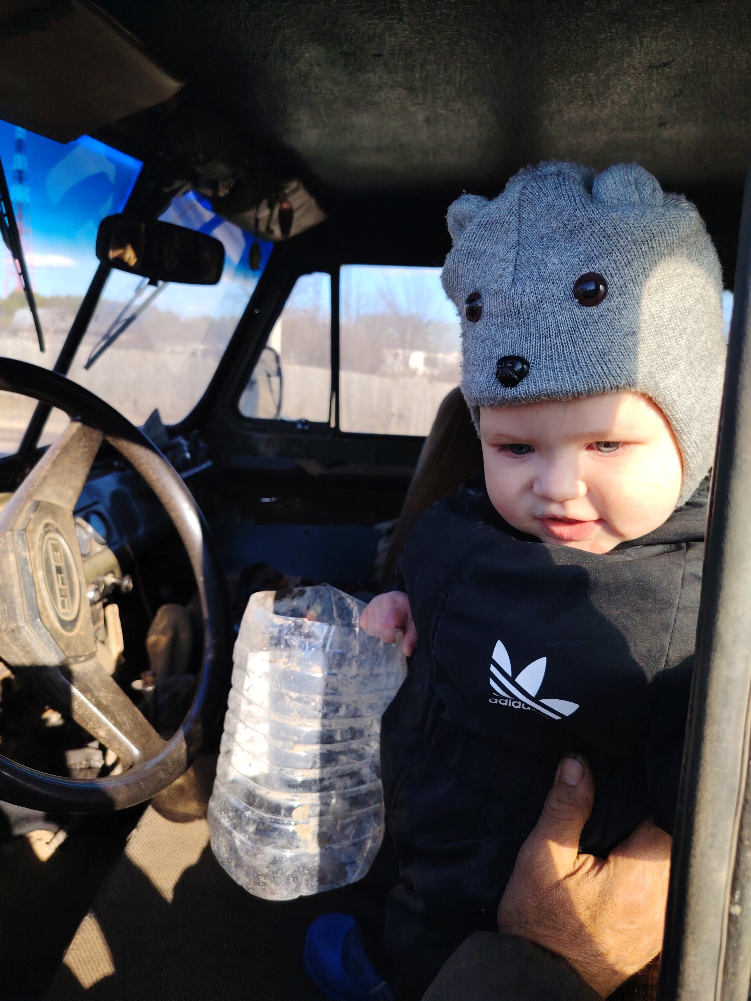 A young child wearing a gray hat with bear-like features sits inside the cabin of what appears to be an old, weathered vehicle, possibly a truck or tractor. The child is wearing a black Adidas jacket and is being held by an adult whose hand is visible. A dirty, clear plastic bottle sits near the steering wheel. The interior of the vehicle looks worn and dusty. The location is possibly Chaikovsky, Russia; however, there is no specific evidence of this in the image itself. Outside the vehicle, through the windshield and side windows, a landscape with vegetation is visible. The lighting suggests it is daytime.