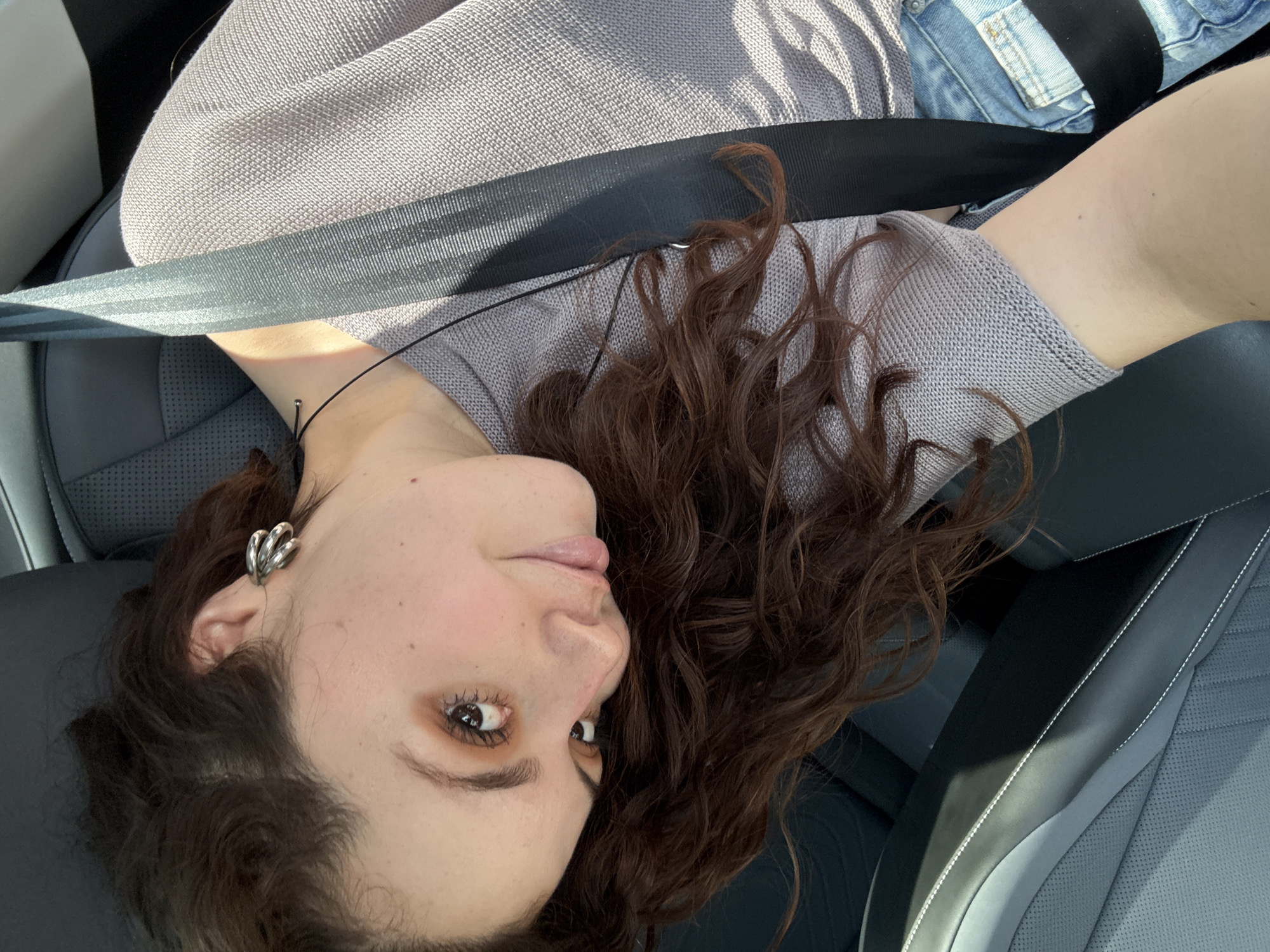 A woman with brown curly hair, wearing a gray sweater and denim shorts, is in the passenger seat of a car. She is wearing a seatbelt and an earring. The car's interior is gray leather. The location is unknown and cannot be determined as Mexico City based on the image.