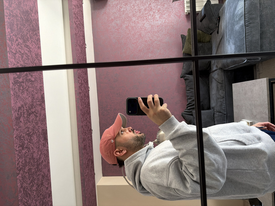 An adult male is depicted taking a mirror selfie in an indoor residential setting within Sofia, Bulgaria. The individual is shown in profile, facing right, wearing a light grey oversized hooded sweatshirt and a faded reddish-orange baseball cap with an emblem. He has facial hair. His right hand holds a black smartphone, positioned to capture his reflection in a multi-panel mirror with dark framing.
The reflection reveals a living area behind the individual. A dark grey sectional sofa with cushions occupies the background, with a remote control visible on one cushion. A light grey rectangular coffee table with a dark metal frame is positioned in the foreground, with a metallic decorative object or lamp base also present on its surface. The walls feature a two-tone design: an upper section with textured, dark magenta wallpaper or paint, separated by a horizontal white band from a lower section of the same dark magenta, extending towards the floor. A white radiator is visible against the far wall. The ceiling is white.
