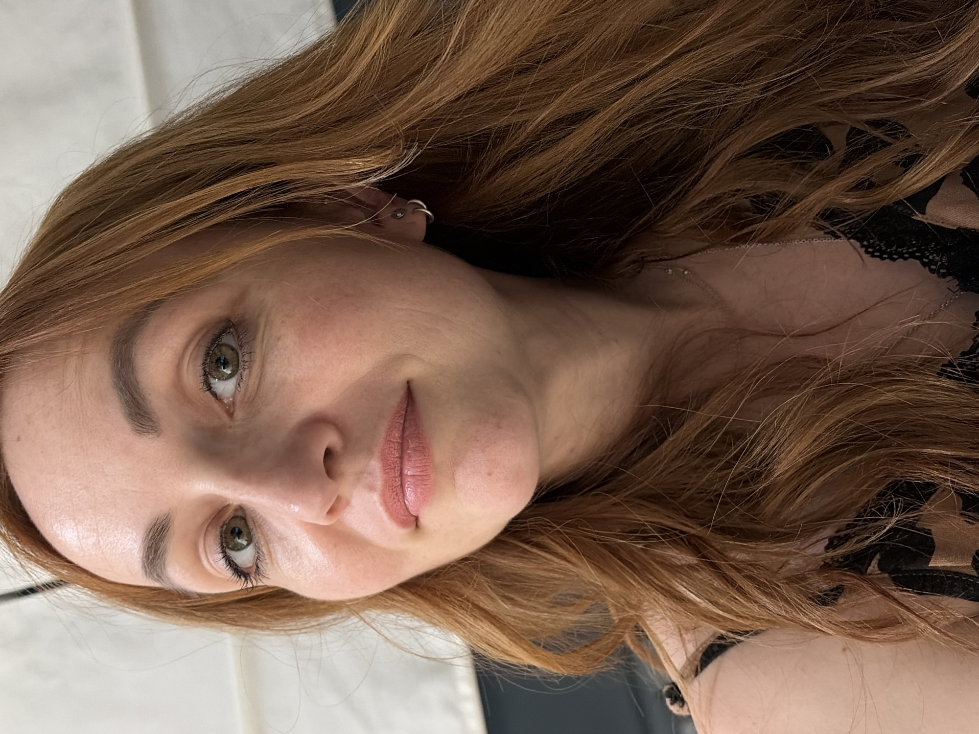 The scene takes place in the city of Chaikovsky, Russia. The image presents a close-up portrait of a woman with long, wavy auburn hair. She has fair skin with a naturally textured complexion, green eyes, and well-defined, filled-in eyebrows. Her eyelashes are coated with mascara, and her lips wear a matte nude-pink lipstick. A faint mole is present on her chin. On her visible left ear, she wears multiple silver earrings, including two hoops and a stud. A thin silver chain necklace is also visible. She is dressed in a top featuring black lace trim and a brown animal print pattern. Her expression is calm with a slight, gentle smile. The background is out of focus, appearing light-colored and indistinct, suggesting an indoor setting.