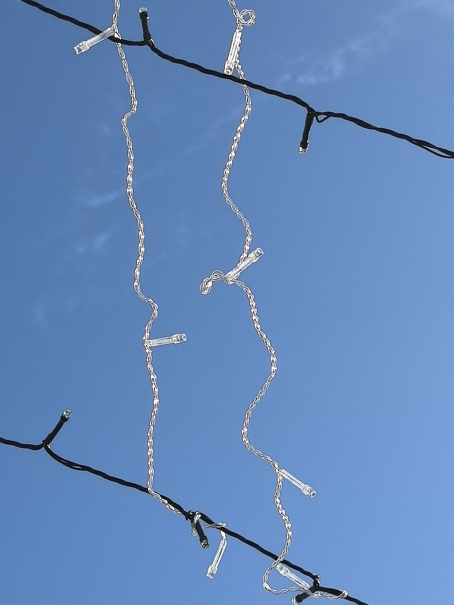 The image captures a close-up, upward view of decorative string lights against a clear blue sky. Two main black electrical wires span the frame horizontally. Attached to these main wires are multiple clear, cylindrical LED light casings. Cascading downwards from the main wires are several distinct strands made of clear, twisted material, each featuring smaller, integrated clear cylindrical light elements along its length, resembling an icicle or fairy light arrangement. The individual light elements are unlit. The background consists of a bright, uniform light blue sky with minimal, faint white wisps of cloud visible in the upper left quadrant. No people or additional structures are present. This scene is located in Noviy, Russia.