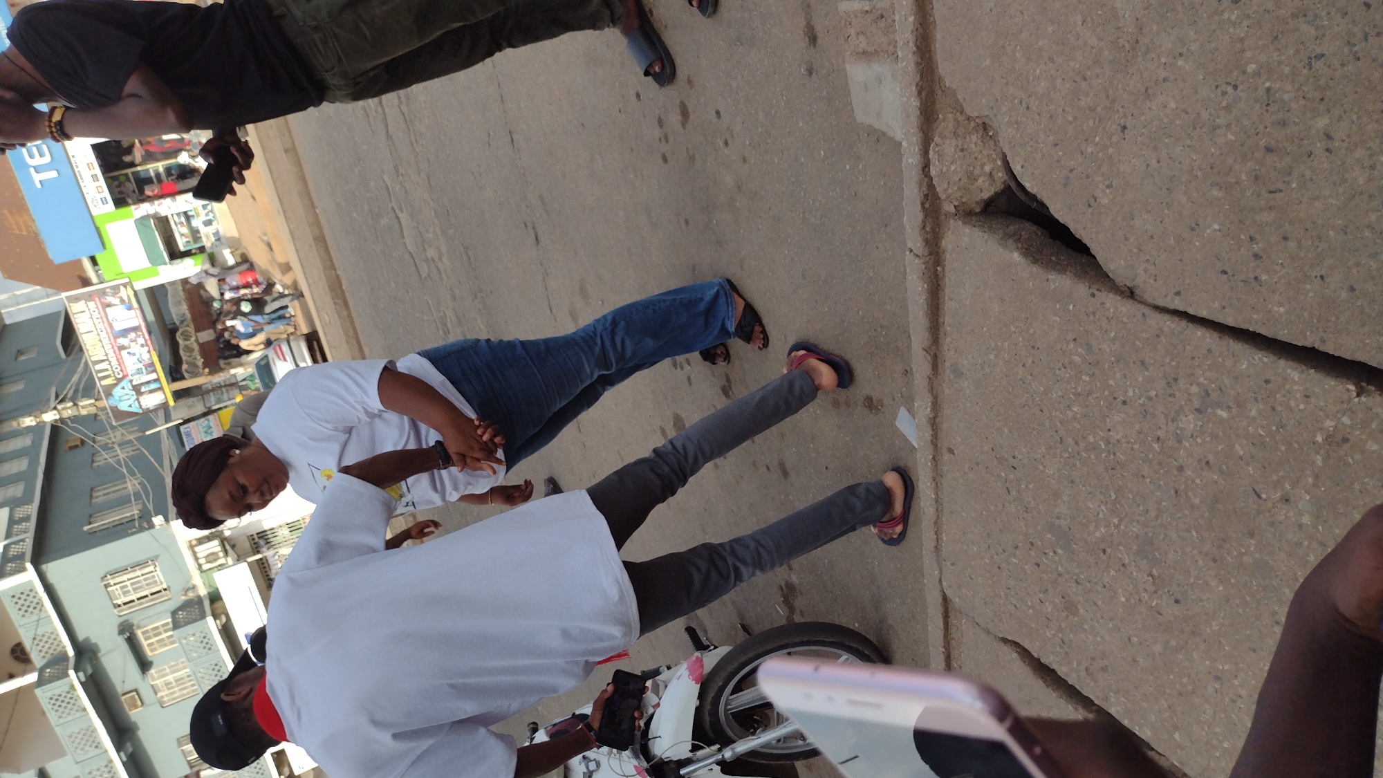 The image depicts a street scene in Jos, Nigeria. Two people are standing on the sidewalk, both wearing white shirts. One person wears blue jeans, and the other wears gray pants. A motorcycle is partially visible between them. To the right, there is a cracked sidewalk and the arm of a person holding a phone taking the picture. A street with buildings on either side can be seen in the background. A sign reading "TEC..." can be seen in the background.