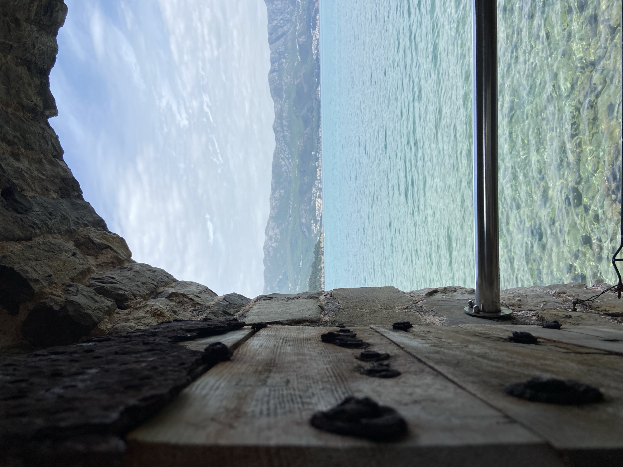 The image, displayed on its side and rotated 90 degrees clockwise, captures a picturesque view from within an old stone structure, looking out over a large body of water toward a distant mountainous coastline. This setting aligns with the historical and natural landscape of Budva, Montenegro.
In the immediate foreground, a weathered wooden surface, possibly a floor or ledge, stretches across the bottom of the frame, featuring several dark, prominent metal studs or fastenings. To the left, rough, dark grey stone walls of the structure frame the view. A shiny, vertical metal pole, perhaps part of a railing or support, rises from the wooden surface on the right side.
Beyond this vantage point lies a vast expanse of calm, turquoise-colored water, its surface gently rippled. Towards the right, the water is remarkably clear, allowing visibility of the bottom beneath. In the midground, a long, rugged mountain range rises steeply from the water's edge, stretching across the horizon. Along the base of these mountains, scattered white buildings indicate a small coastal settlement. The sky above is a bright, clear blue, adorned with soft, wispy white clouds.
The scene depicts a tranquil daytime moment, likely morning or early afternoon, given the bright yet soft light. No specific activities or events are occurring, nor are there any people or visible text in the image. The composition evokes a sense of historical grandeur combined with natural beauty.