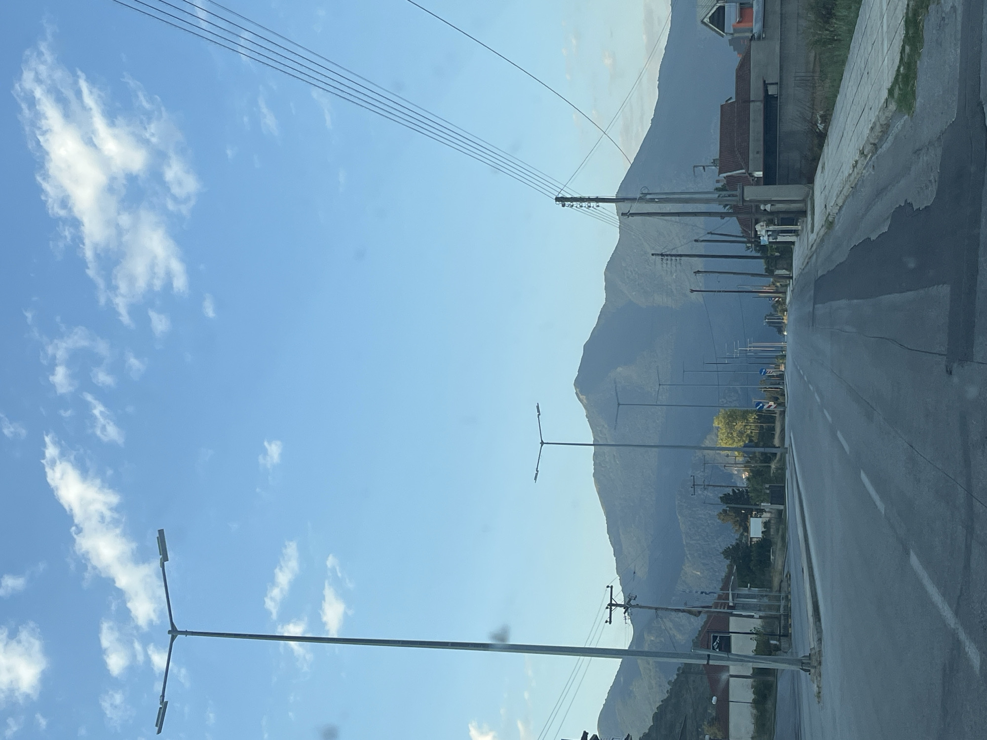 The image depicts a street scene in Kato Nevrokopi, Greece, with a paved road leading towards a large, tree-covered mountain under a bright blue sky with scattered white clouds. On the left side of the road, a tall lamppost with two lights is visible, along with power poles and wires. Buildings with red-tiled roofs and some residential structures are present on the right side of the road. Several other lampposts are spaced along the road, and a few signs are visible in the distance. The scene appears to be a quiet, daytime view of a town approaching mountainous terrain.