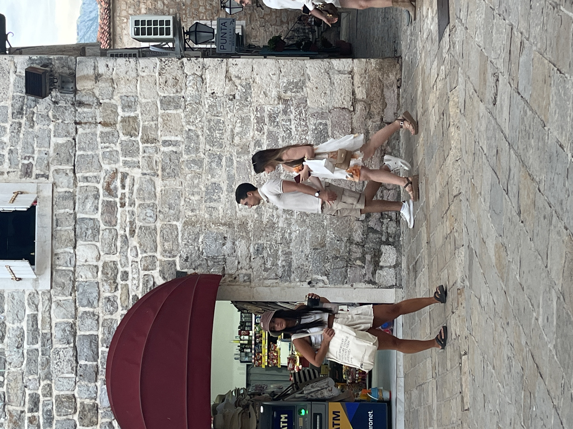 A woman walks down a cobblestone street in Budva, Montenegro, carrying a tote bag and a cup. She is wearing a white tank top, a bucket hat, and light-colored shorts. Behind her, a couple walks past, the man in a light-colored polo shirt and shorts, and the woman in a white floral dress. A building with a stone facade and a red awning is on the left, and a sign for "PUNTA KOMORA RESTAURANT" is visible on the right. The scene appears to be set in a historic part of the city, likely in a pedestrian area.