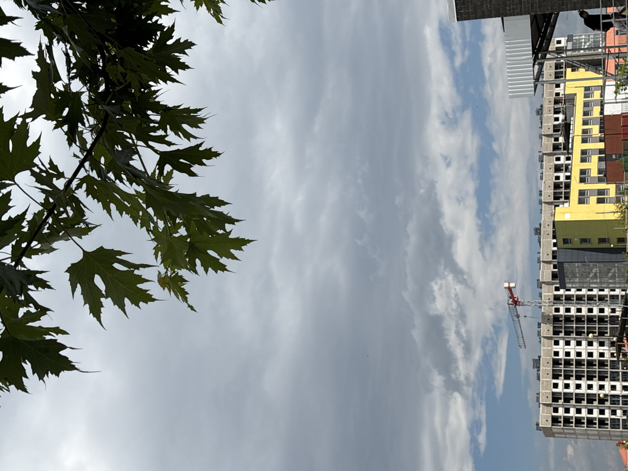 The image depicts a construction site in Mikhaylovsk, Russia, on a cloudy day. In the foreground, green leaves hang from a tree branch, obscuring the upper portion of the frame. Below, a large, multi-story building under construction dominates the middle ground. The building has a repetitive pattern of open window frames. A red and white construction crane is visible to the left of the building. Adjacent to the unfinished building, a brightly colored yellow structure with multiple windows is visible. In the right portion of the image, a person in dark clothing stands near a portion of a building with dark brickwork and a white, ribbed structure extending outwards. The sky is filled with a mix of gray clouds and patches of blue sky.