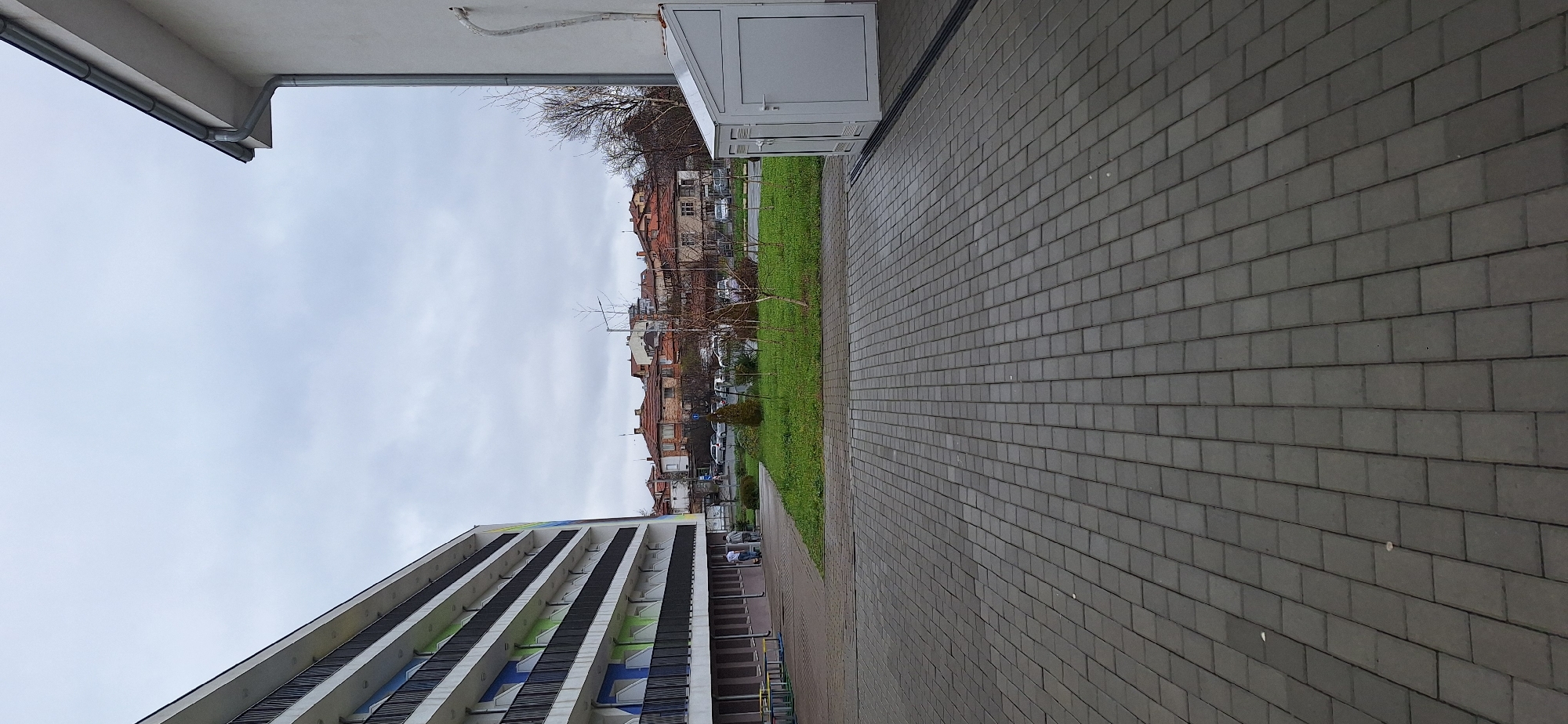 The image shows an outdoor scene in Burgas, Bulgaria. In the foreground is a paved gray brick walkway on the right and a strip of green grass and sparsely planted trees in the center. To the left is a tall building with horizontal lines of windows and balconies. Across the green strip are residential buildings with red tile roofs. The sky is overcast.
