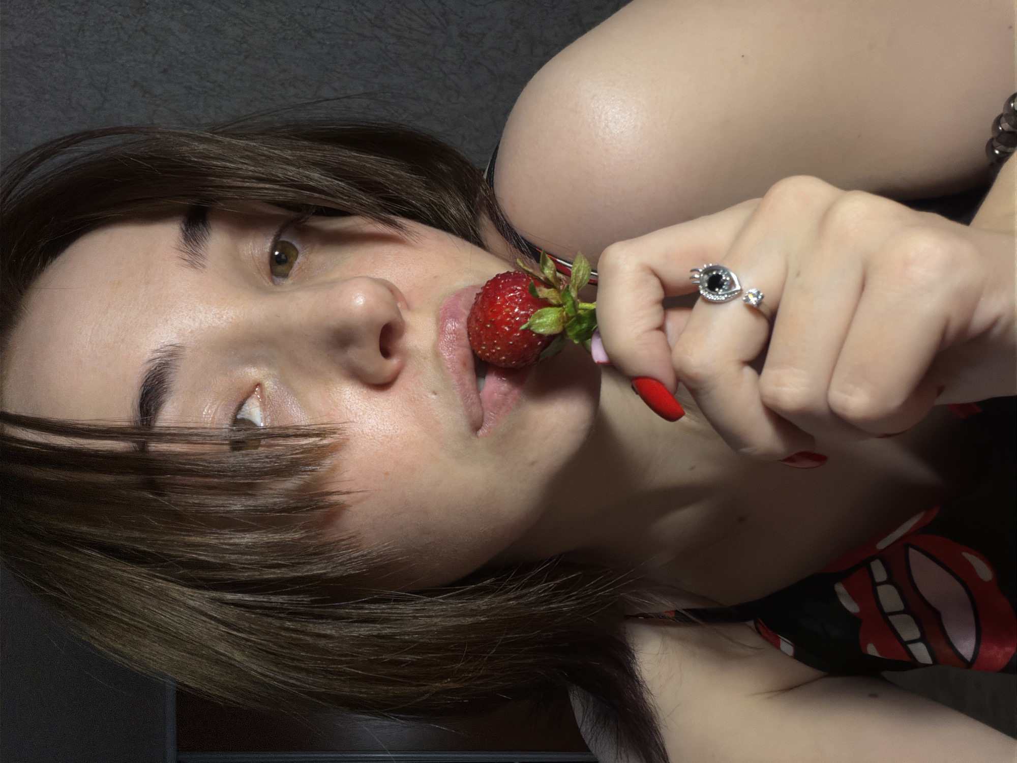 An unknown young woman is captured in a close-up shot, holding a strawberry to her mouth. Her dark hair is styled with bangs that partially obscure her face. She is wearing a black top with a bold red lips and teeth pattern. Her right hand, adorned with bright red nail polish and a ring with a black stone and a smaller diamond, is holding the strawberry near her lips, which are slightly parted as if about to take a bite. The background is a plain, dark textured wall. The lighting suggests an indoor setting, possibly a home environment.