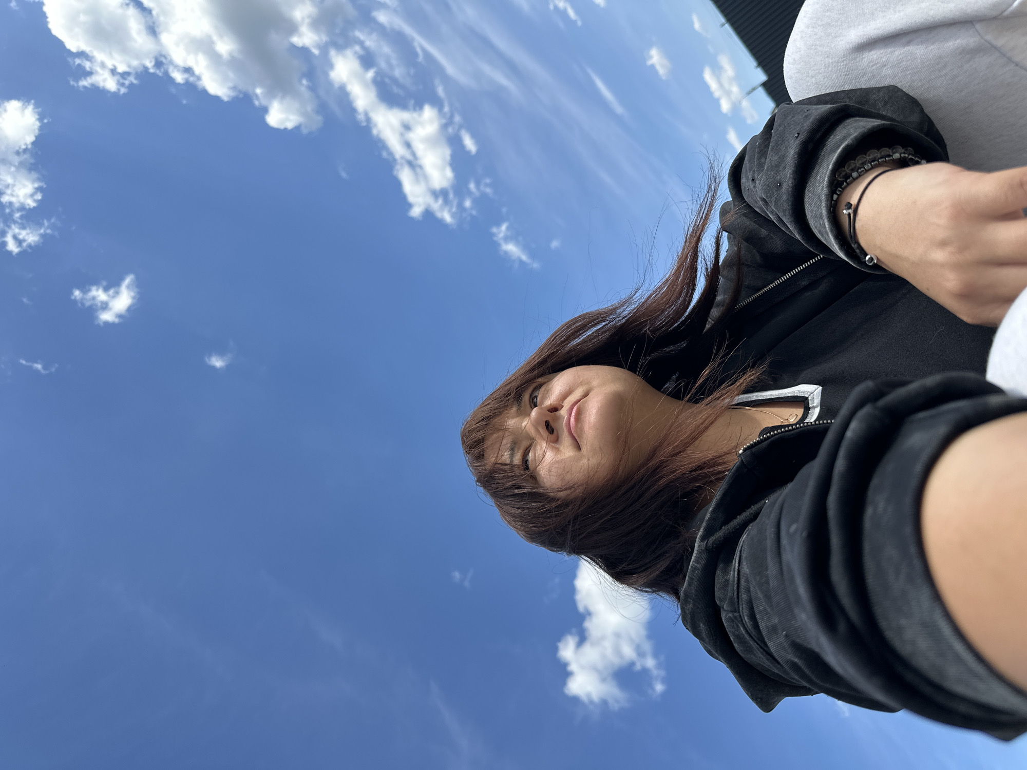 A low-angle, eye-level shot captures a woman against a backdrop of a blue sky speckled with white clouds. She is wearing a black hoodie with a white stripe near the zipper, and dark bracelets adorn her wrist. Her brown hair is windswept. The location is Unknown, Unknown.