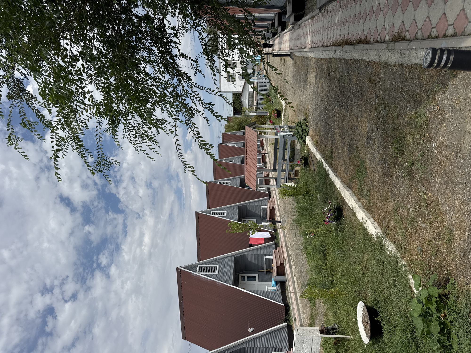 In Zharbulak, Kazakhstan, a row of A-frame cabins with dark red roofs and grey siding is visible. The cabins are lined up along a gravel path. A paved walkway runs alongside the cabins on the right side of the image. There is vegetation and scattered flowers around the cabins. A person is walking on the paved walkway in the distance. The sky is cloudy.