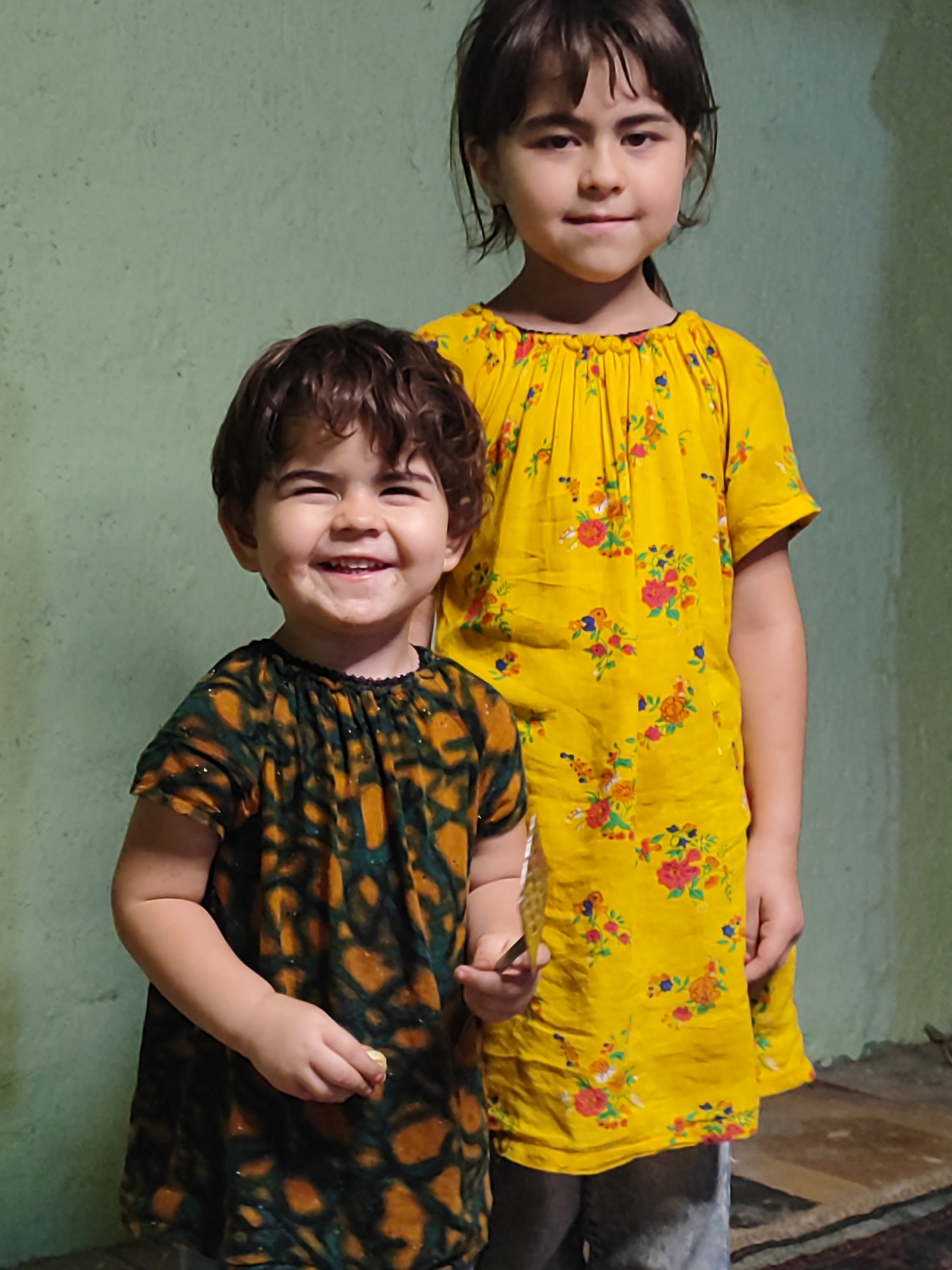 Two young girls, appearing to be of Central Asian descent, are standing in front of a light green textured wall. The girl in the foreground, younger and on the left, has curly brown hair and is wearing a dark green and orange patterned dress. She is smiling broadly and holding a small object, possibly food, in her right hand. Behind her and slightly to the right stands an older girl with dark brown hair, wearing a bright yellow dress adorned with a floral print in shades of red, blue, and orange. Both girls are facing forward, and the older girl has a slight smile. The environment suggests an indoor setting, possibly a home, with a glimpse of a rug on the floor to the right. The overall scene is brightly lit. Based on the provided information, this image is not associated with the city of Ismoili Somoni, Tajikistan, as no specific location details were provided beyond the image itself.