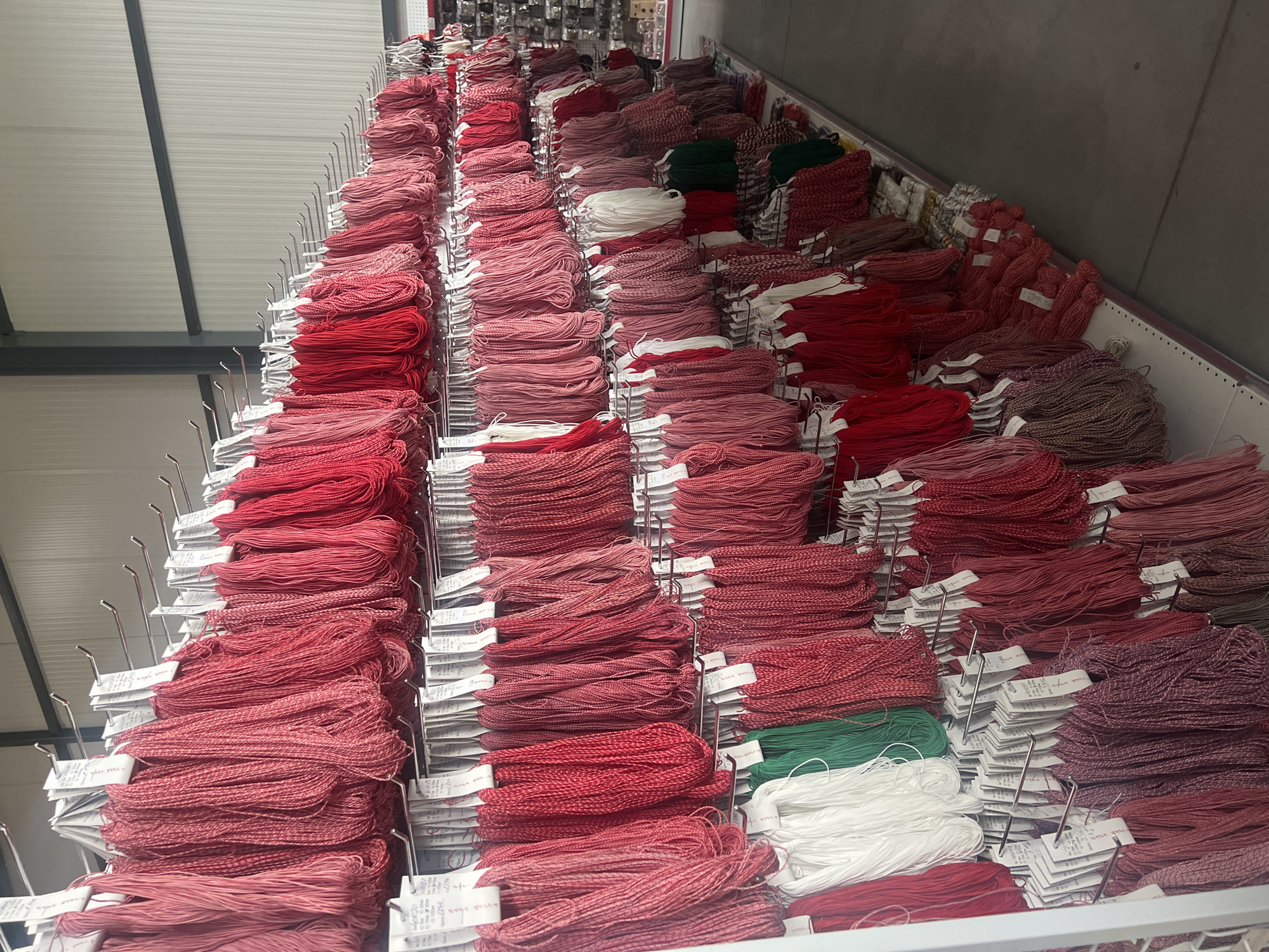 The image depicts a warehouse-like setting in Burgas, Bulgaria, filled with rows of neatly stacked bundles of embroidery floss or thread. The dominant colors are shades of red, ranging from bright crimson to muted pink and maroon, with a few bundles of white and green floss interspersed. The floss is arranged on metal racks with hanging arms, each bundle accompanied by a small white tag, presumably containing information about the thread type, color code, or price. The environment appears industrial, with overhead metal beams and a corrugated ceiling. The focus is on the vast quantity of thread, indicating a store or storage facility dedicated to crafting supplies.