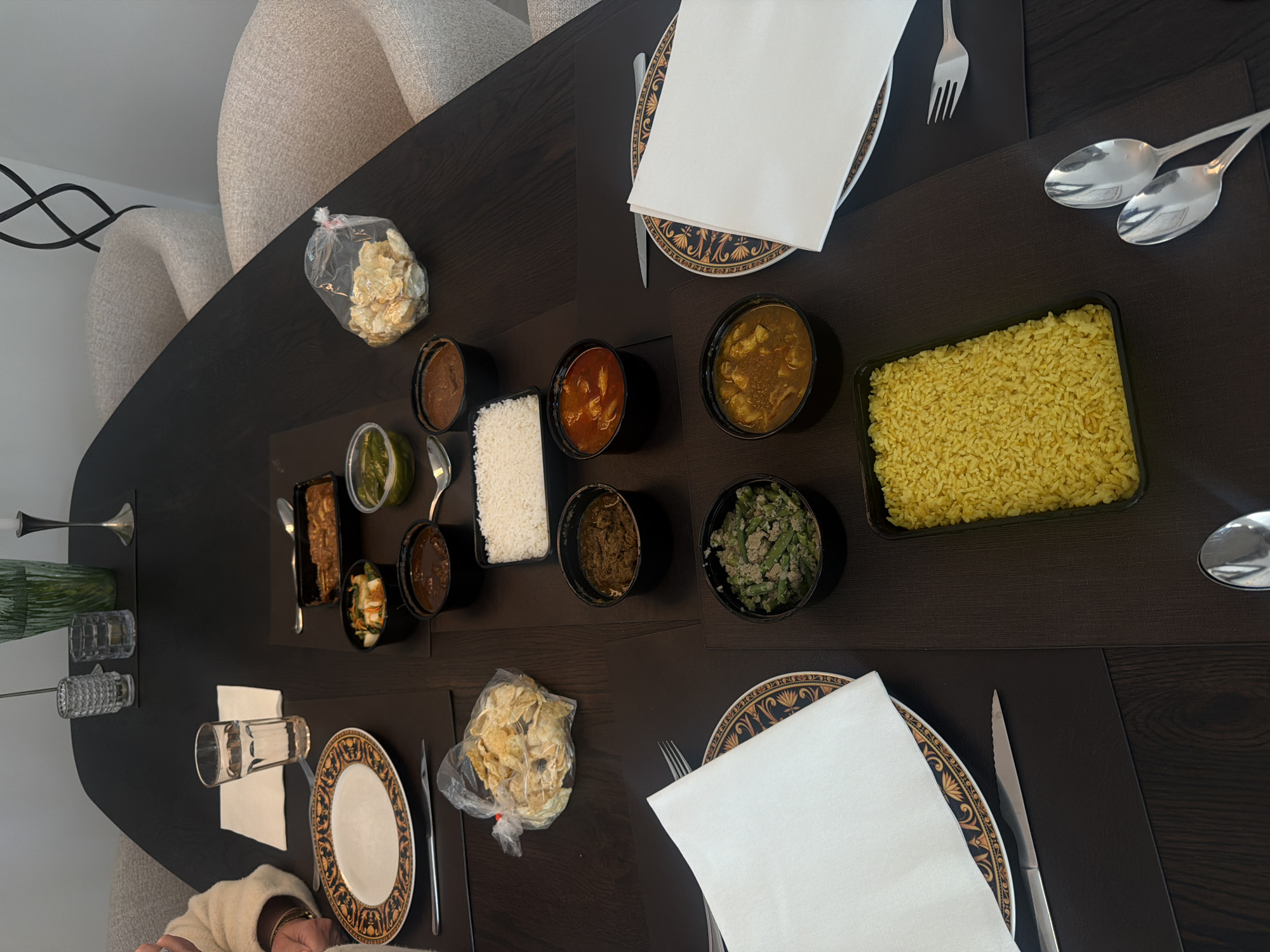 The image depicts a dining table set for a meal, likely in Ridderkerk, Netherlands, as inferred from the context. The table is dark and appears to be made of wood. Several place settings are visible, each with a plate, napkin, and cutlery. The plates have an ornate, gold and black pattern.
Various dishes are arranged on the table, suggesting a meal, possibly Indian or South Asian cuisine. These include containers of rice (both white and yellow), curries or stews in black bowls, a salad or vegetable dish in a black bowl, and a bag of what appear to be puffed snacks or papadums. There are also small containers with a green sauce or relish. A glass of water and two decorative glasses are also present on the table.
In the background, two upholstered chairs are visible, suggesting a home or restaurant setting. A person's arm and shoulder are visible on the left side of the frame, indicating their presence at the table. The overall lighting is somewhat dim, with light sources not directly visible but contributing to a warm ambiance. The scene is static, with no apparent action occurring.