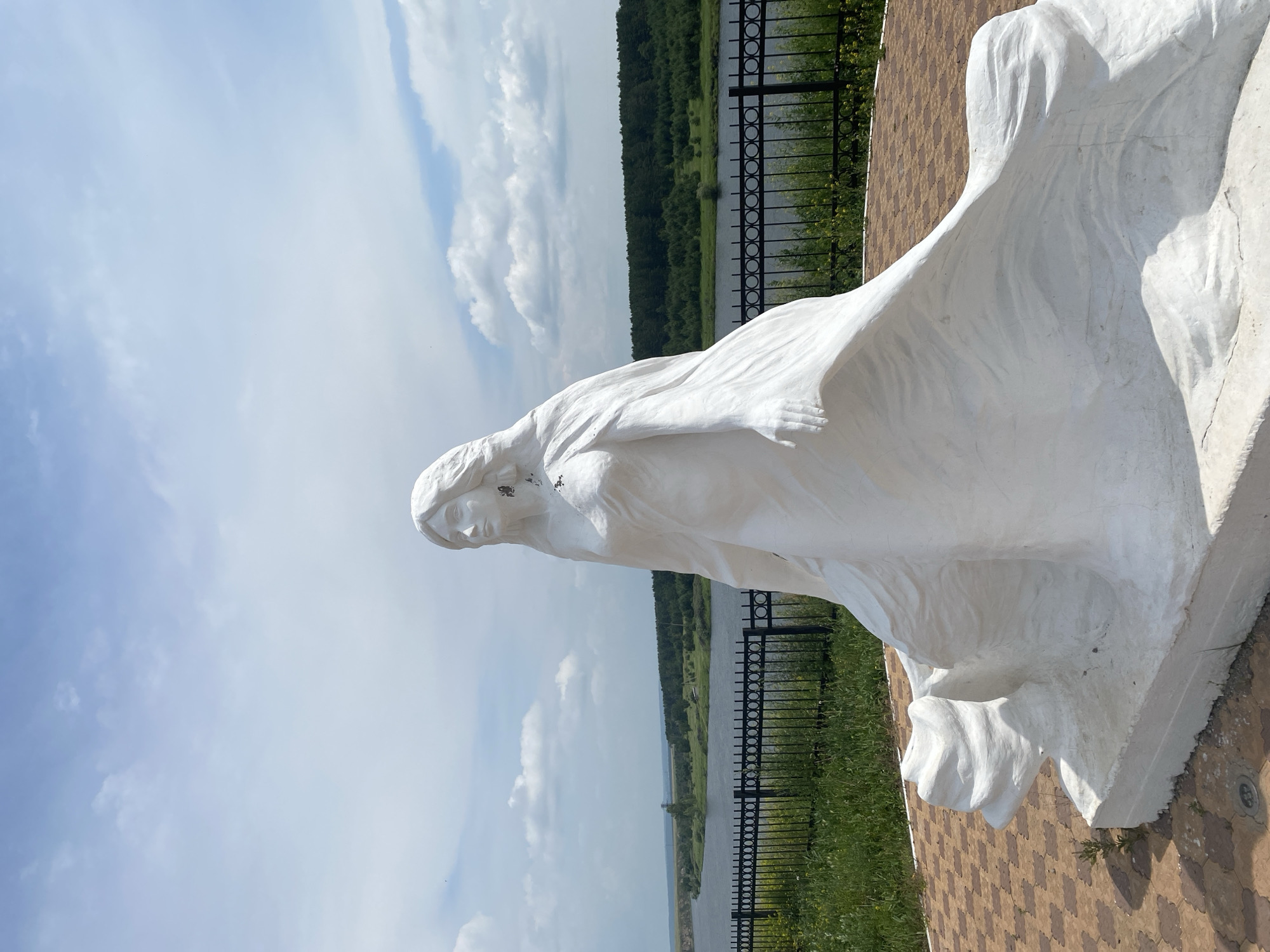 A white stone sculpture of a woman stands in front of a river and a treeline under a cloudy sky. The sculpture is depicted in a flowing gown, with her hair falling around her shoulders. The woman's head is turned to the right, and her expression is serene. The sculpture is positioned on a raised circular platform paved with brown and beige interlocking tiles. A black metal fence runs along the edge of the platform, separating it from a grassy area with yellow wildflowers. The river in the background is wide and calm, reflecting the sky. Beyond the river, a dense forest of green trees stretches across the horizon. The sky is blue with scattered white clouds, suggesting a pleasant day. The location is specified as Novomaltinsk, Russia.