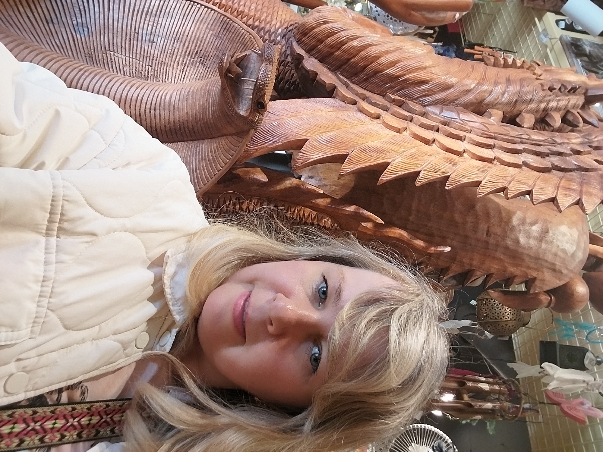 A young woman with blonde hair and blue eyes is captured in a close-up, slightly angled selfie, offering a gentle smile. She is wearing a light-colored, possibly cream or off-white, quilted jacket. Her expression conveys a sense of mild pleasantness or contentment.
The most prominent background element is a large, intricately carved wooden sculpture. Made from a dark, rich wood, the carving features detailed textures resembling feathers, scales, and a formidable head with an eye and an open mouth. It appears to depict a mythical creature, possibly a dragon or a large bird, showcasing complex craftsmanship.
The setting appears to be indoors, likely a shop, market, or exhibition space in Sochi, Russia, specializing in artisan goods or decorative items. Hints of other objects are visible in the background, including a patterned fabric border at the bottom left, a circular fan-like object, and various decorative pieces such as a small, ornate metal lantern with geometric cutouts, and other miscellaneous items on what looks like a display grid. The woman's pose suggests she is taking a selfie or being photographed, highlighting both herself and the impressive wooden artwork.